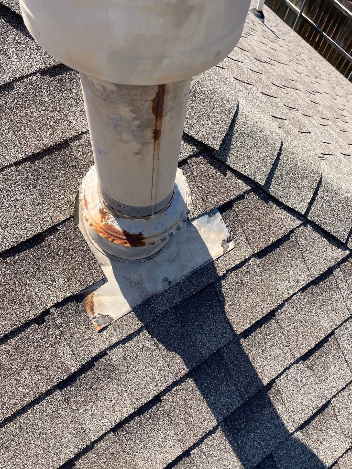 Close-up of a rusted metal vent pipe protruding from a shingled roof with visible rust stains and shadow.