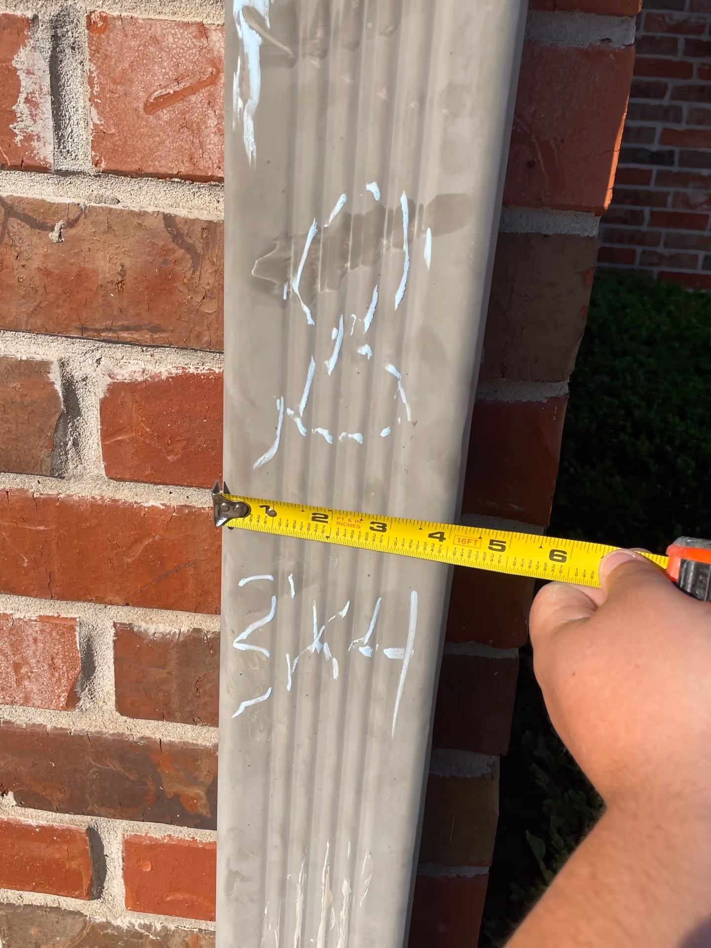 Hand holding a yellow measuring tape showing a width of about 5 inches against a beige downspout with white chalk markings, set against a red brick wall.