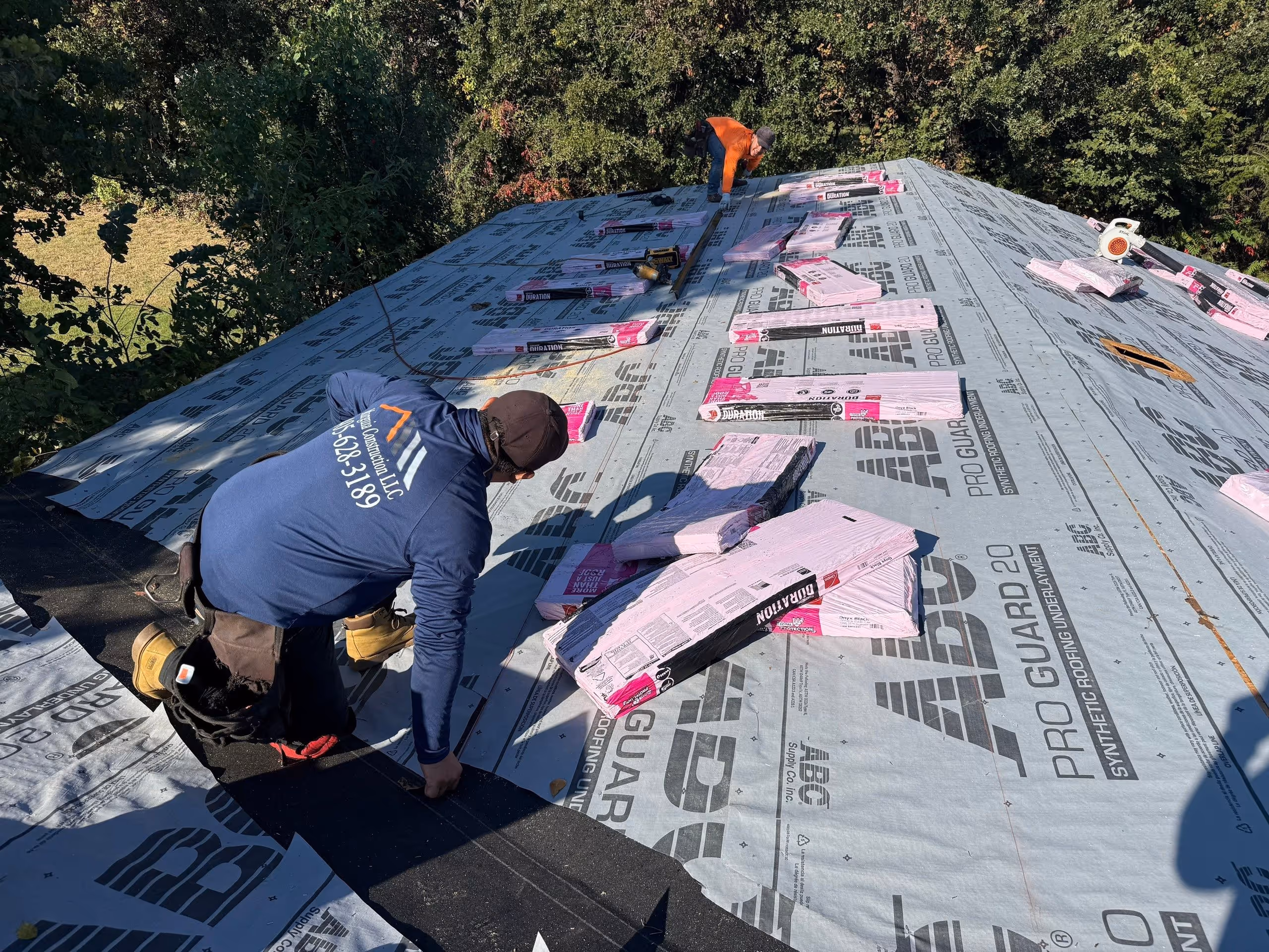 Two roofers installing synthetic roofing underlayment on a sloped roof surrounded by trees.
