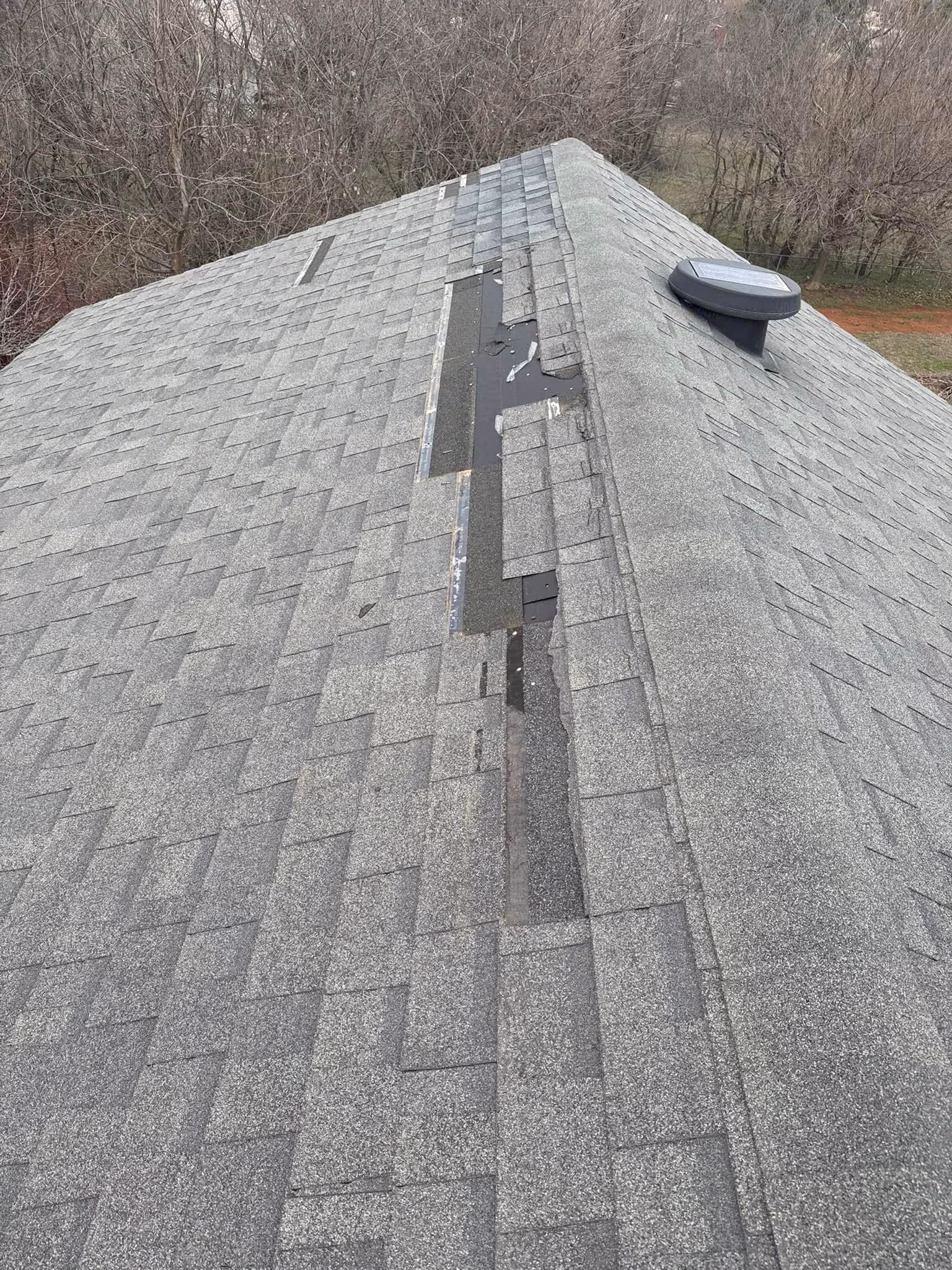 Gray shingled roof with a long strip of missing shingles revealing black underlayment near the ridge vent after a recent Oklahoma City storm.