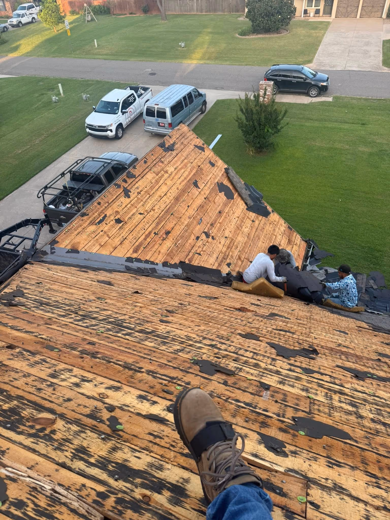 View from a rooftop showing workers removing old shingles, exposing wooden roof decking; parked trucks and green lawns visible below.