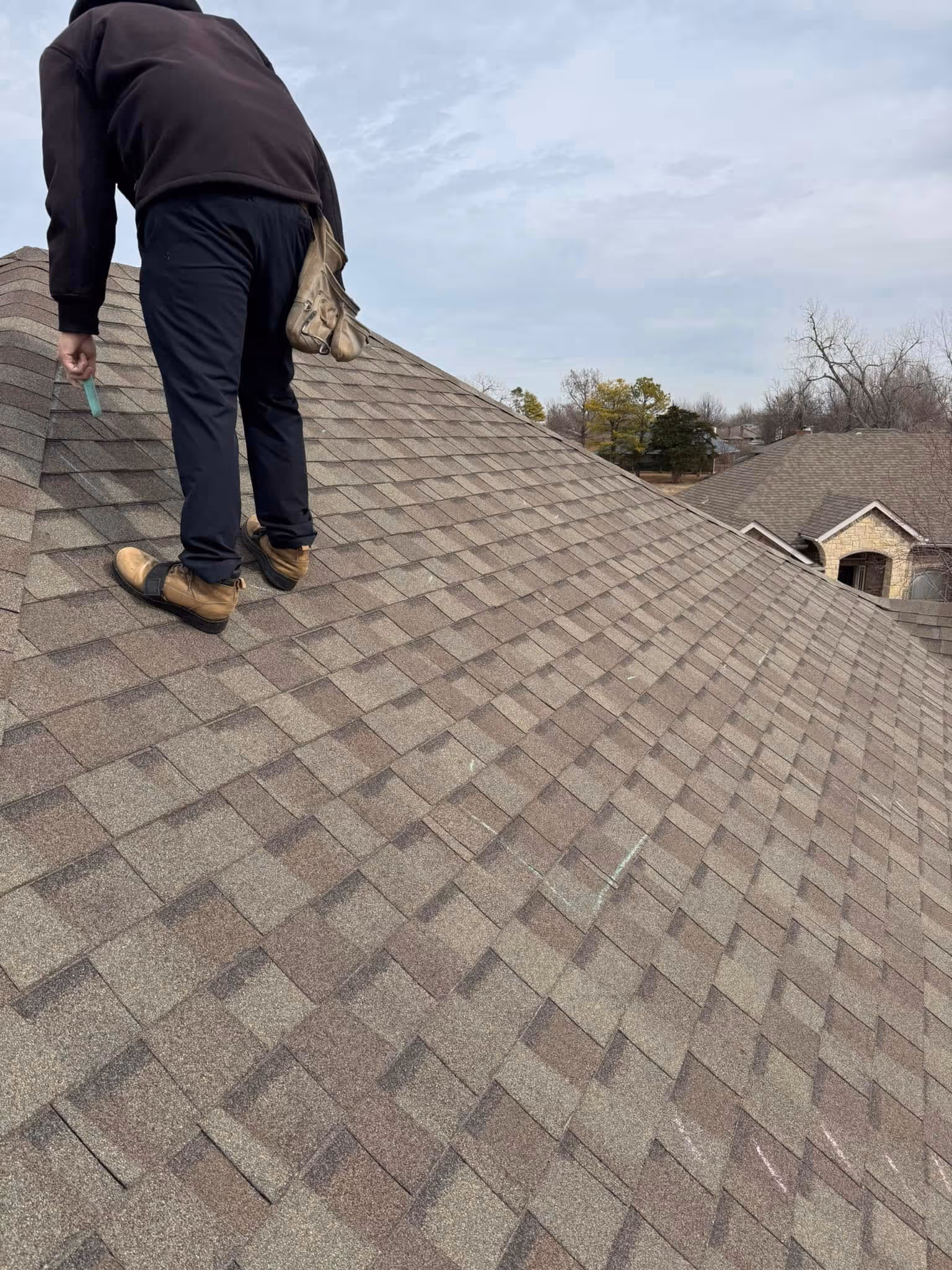 Person wearing work boots and black pants inspecting and marking damaged shingles on a brown shingled roof under a cloudy Oklahoma sky.