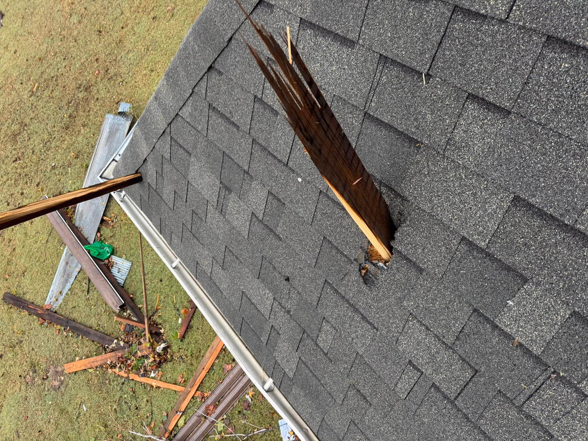 Close-up of an Oklahoma storm damaged shingle roof with a large splintered wooden pole pierced through it, debris scattered on grass below.