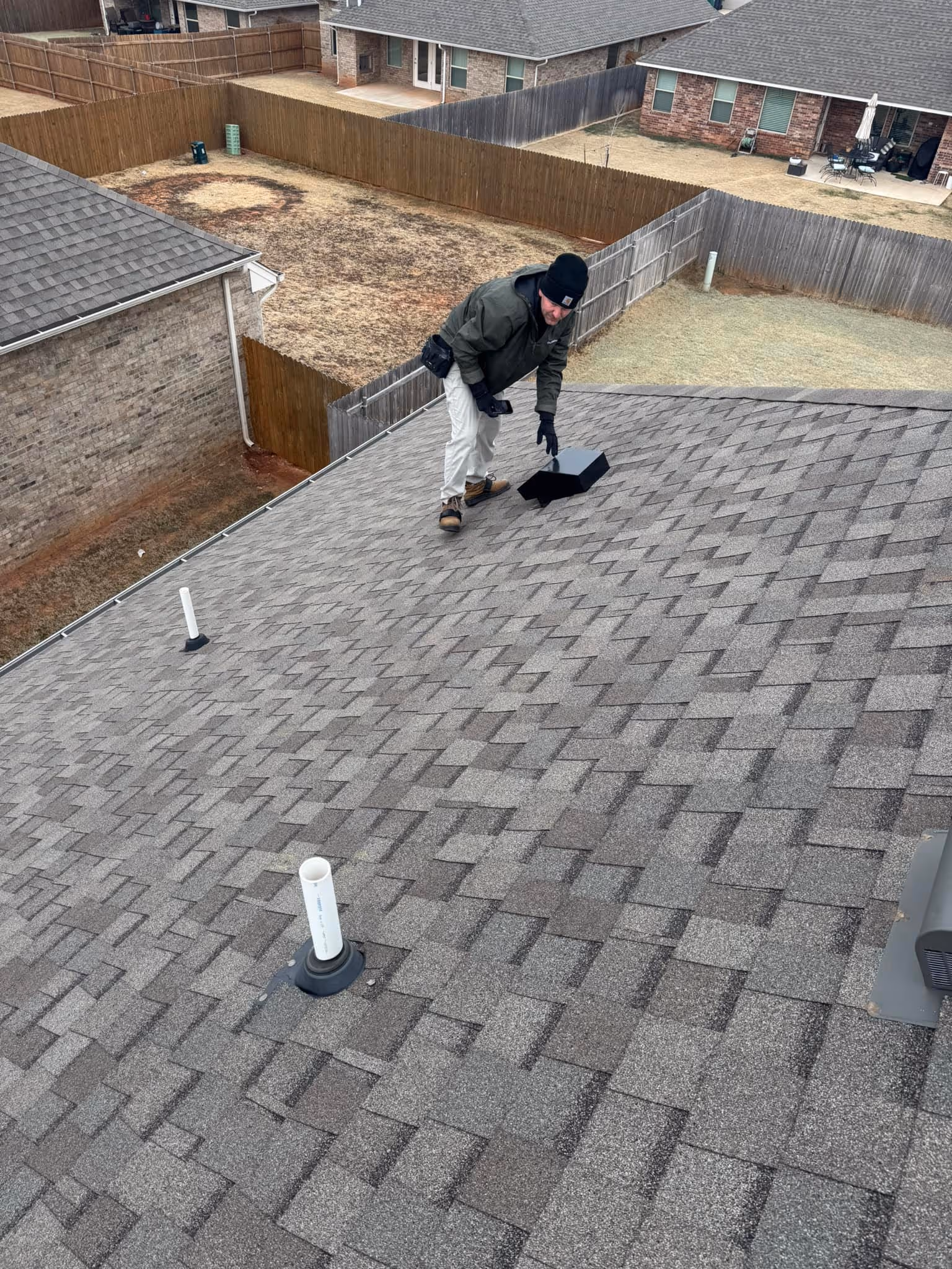 Man wearing gloves and a jacket working on a gray shingled roof near vents and a black roof vent cover.