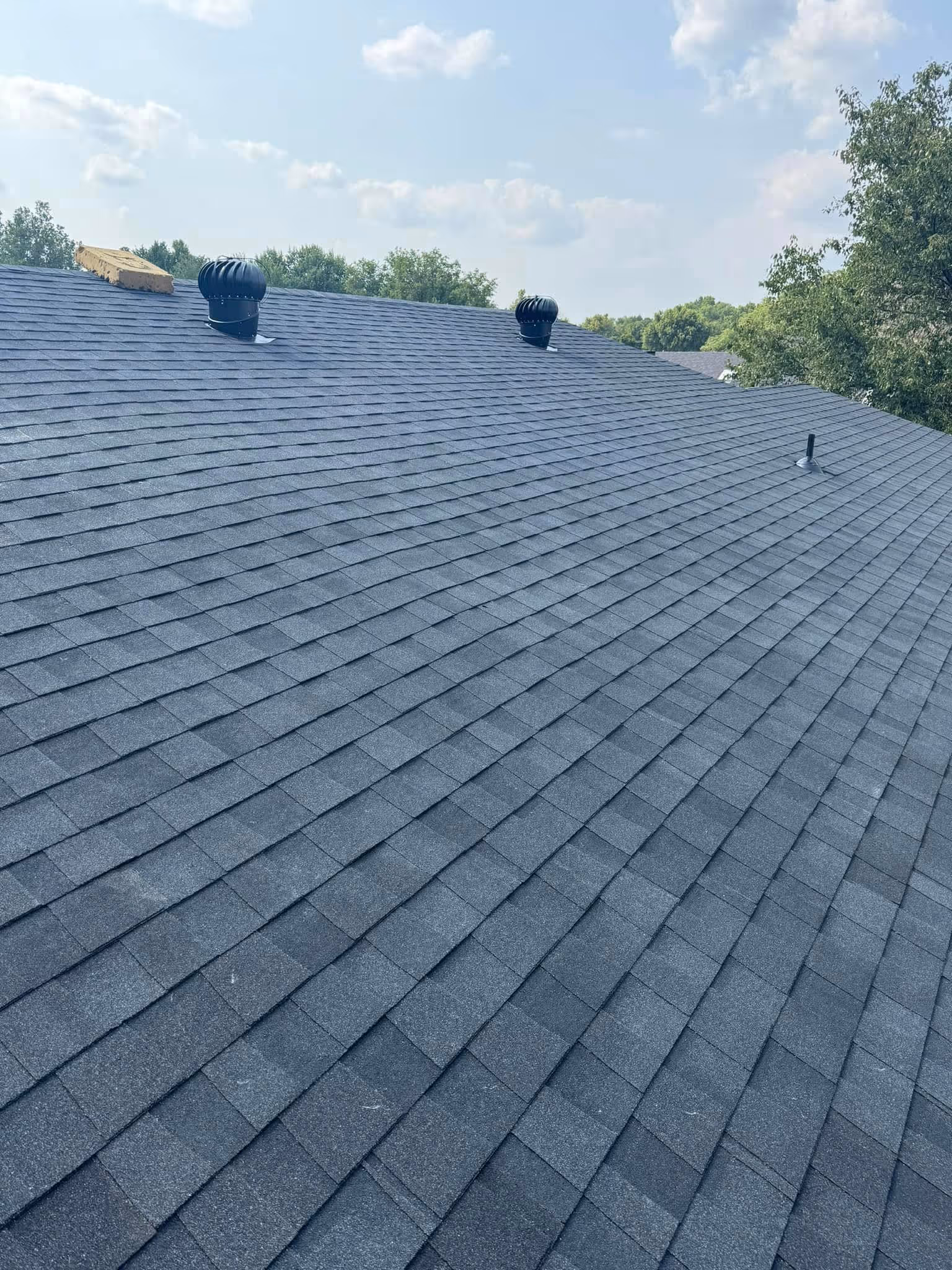Newly installed dark gray asphalt shingle roof with two black roof vents and trees in the background under a partly cloudy sky.
