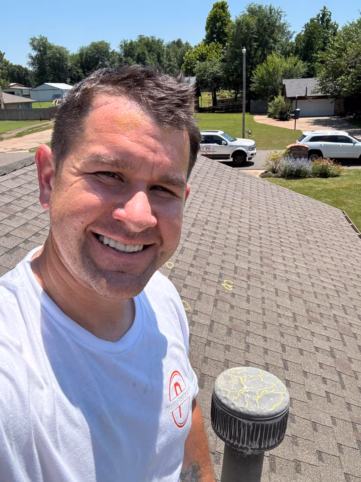 Smiling man in a white shirt standing on a roof with asphalt shingles and a vent pipe marked with yellow chalk.