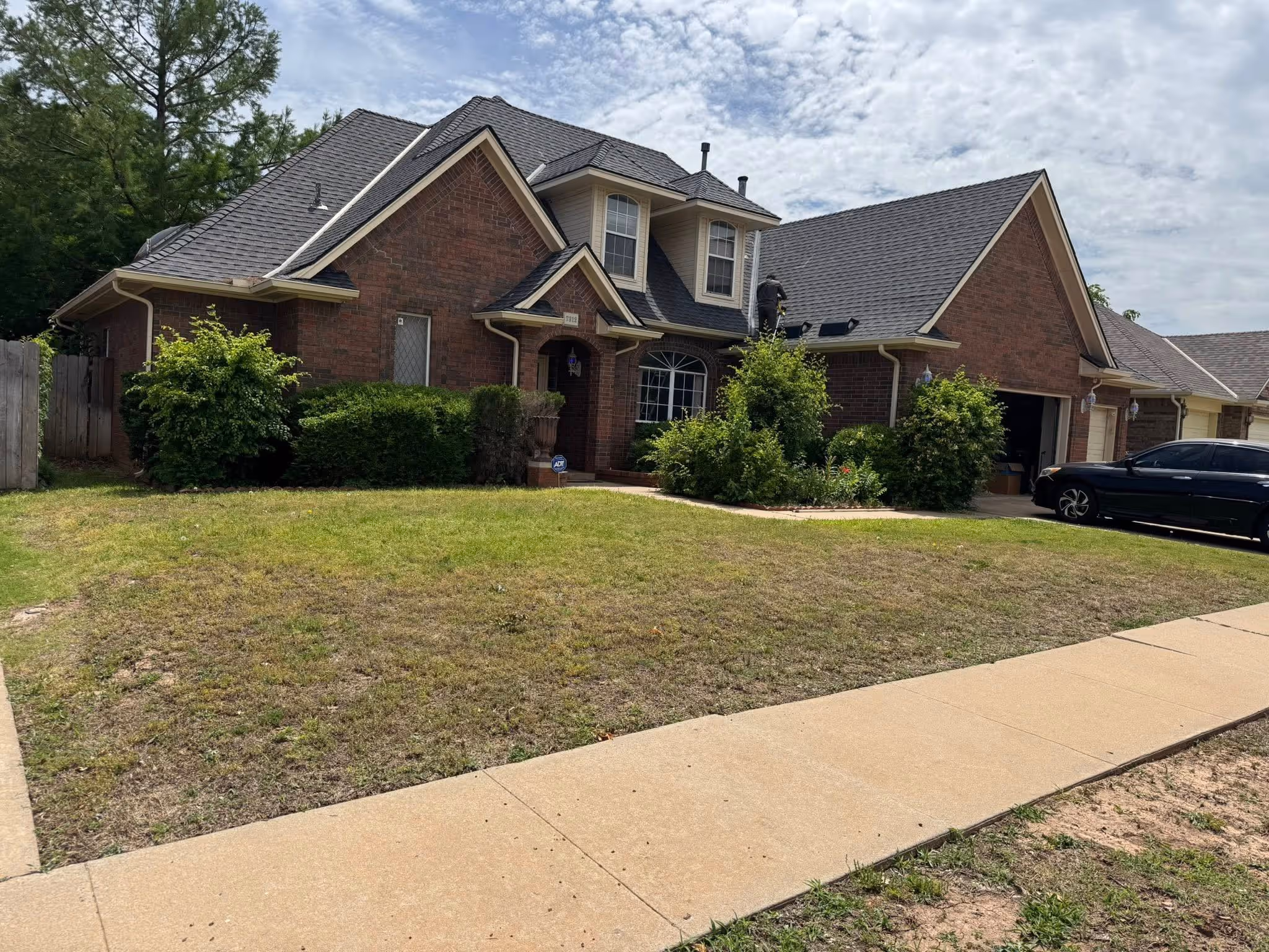 Brick house with dark gray roof and a person working on the roof next to a green lawn and driveway with a black car parked.
