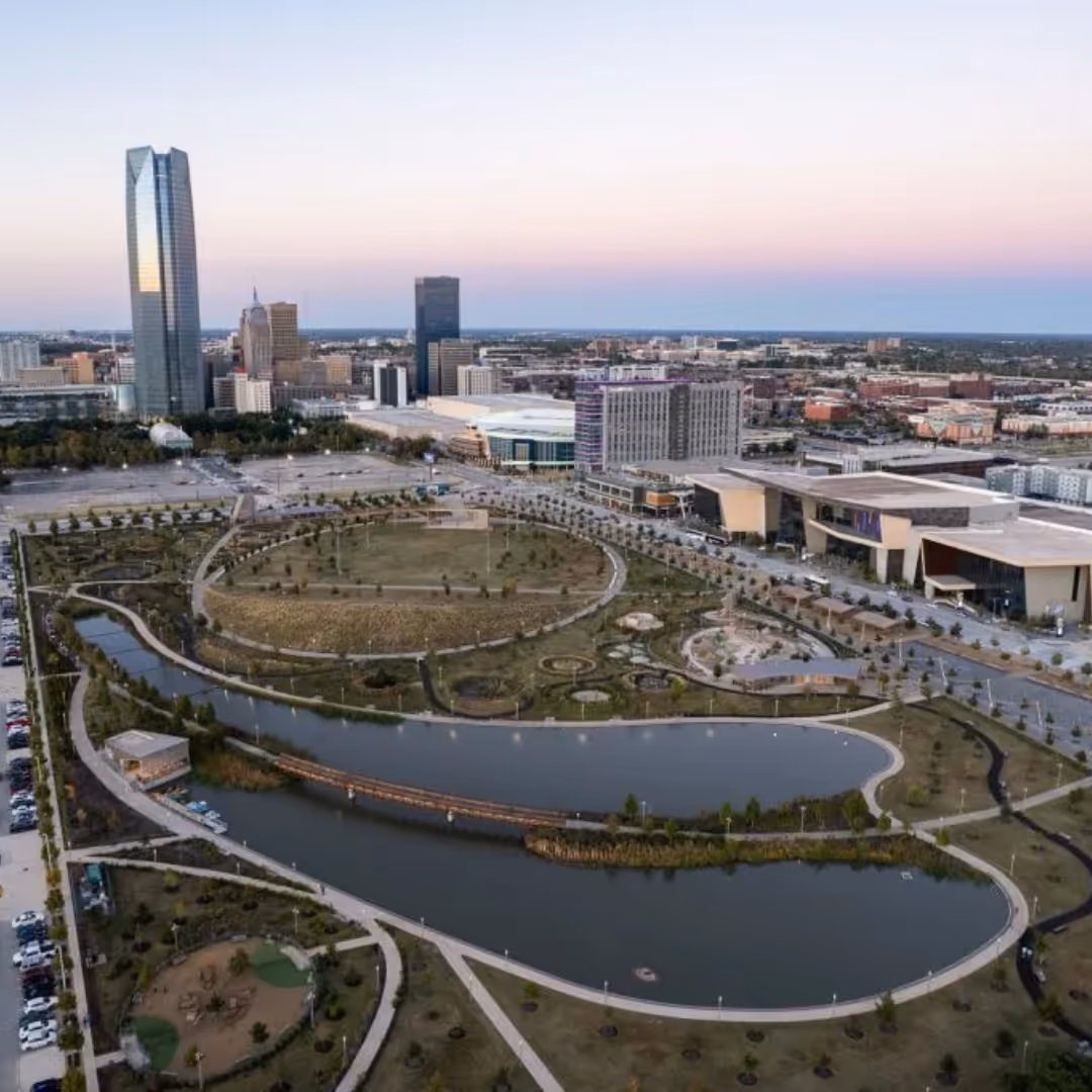 Aerial view of the Oklahoma City Scissortail park with winding waterways and trails, surrounded by urban buildings and a tall skyscraper at sunset.