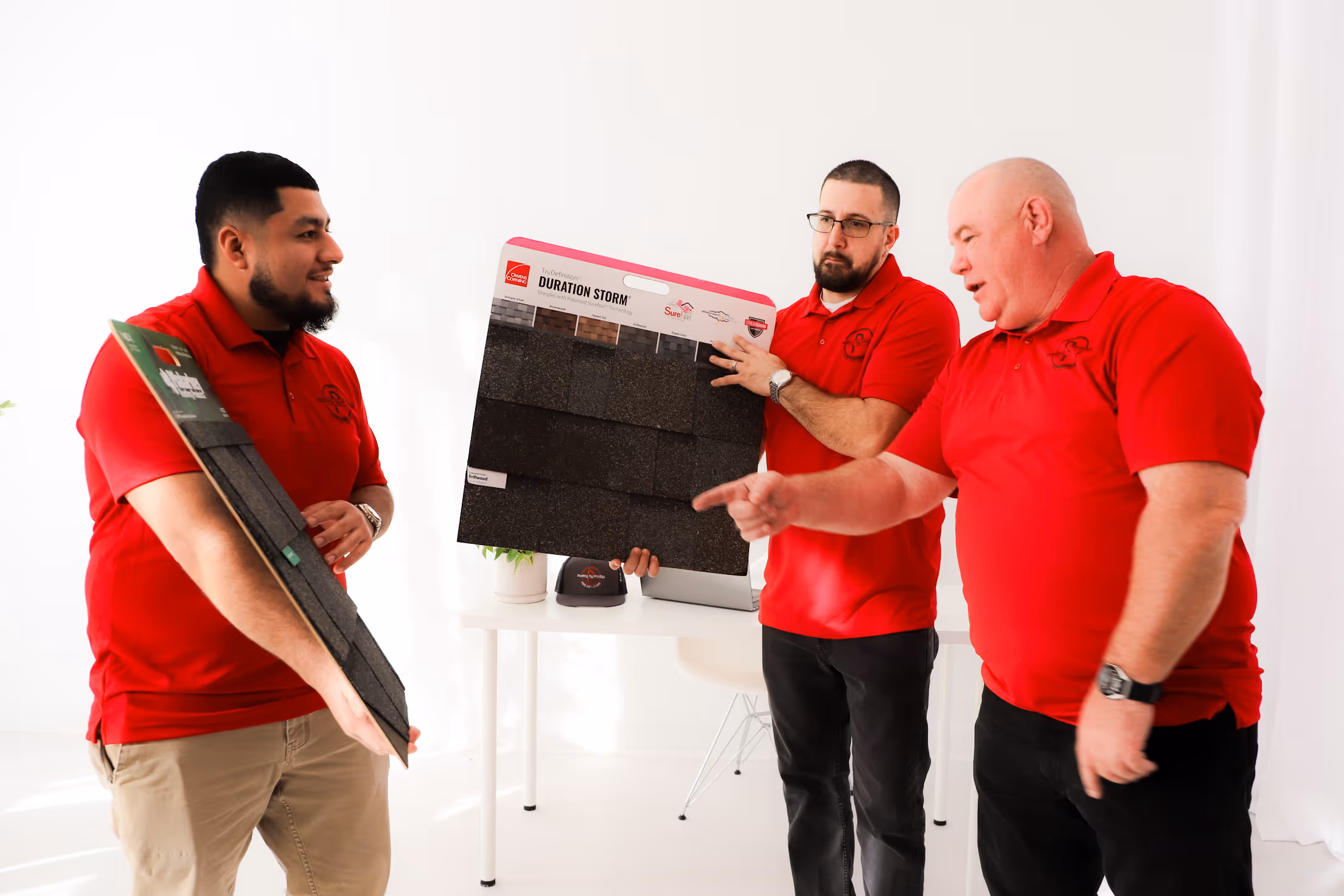 Roofing by Prodigy - Three men in red polo shirts discussing and holding roofing shingle samples in a bright room.