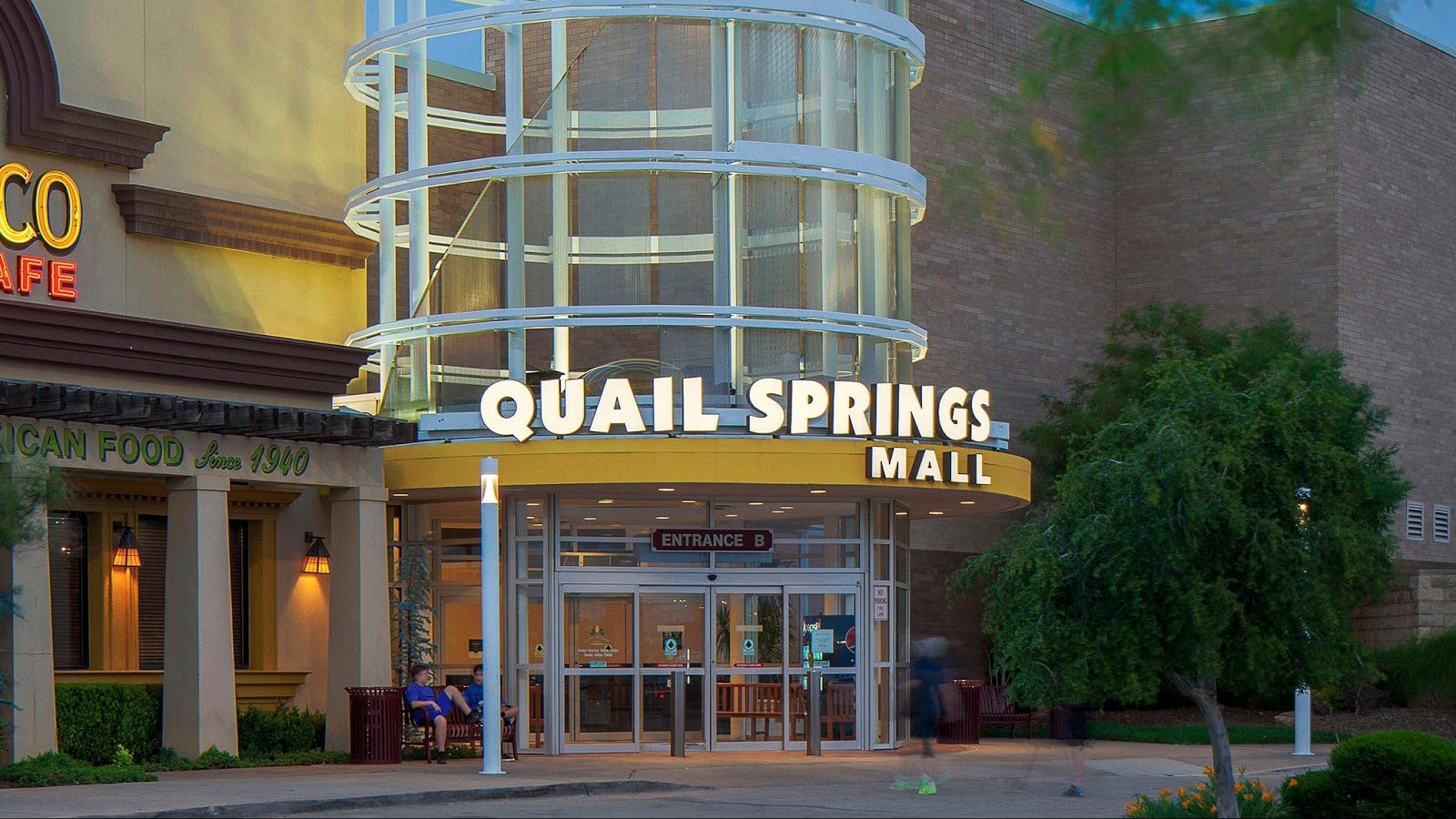 Entrance B of Quail Springs Mall with illuminated mall sign, adjacent restaurant, and people sitting and walking.