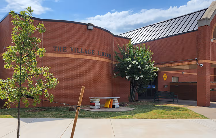 Exterior of The Village Library building with red brick walls, a tree, a bench shaped like stacked books, and a clear blue sky.