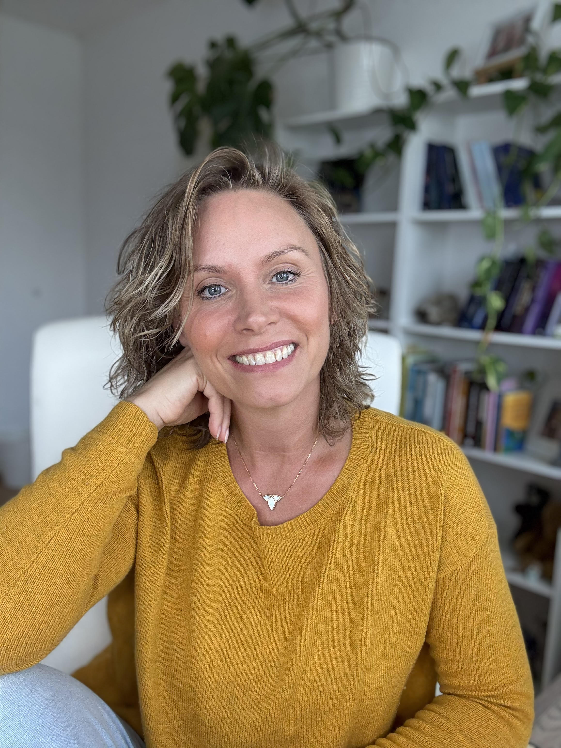 Frau mit lockigem Haar in gelbem Pullover lächelt entspannt vor einem Bücherregal mit Pflanzen.