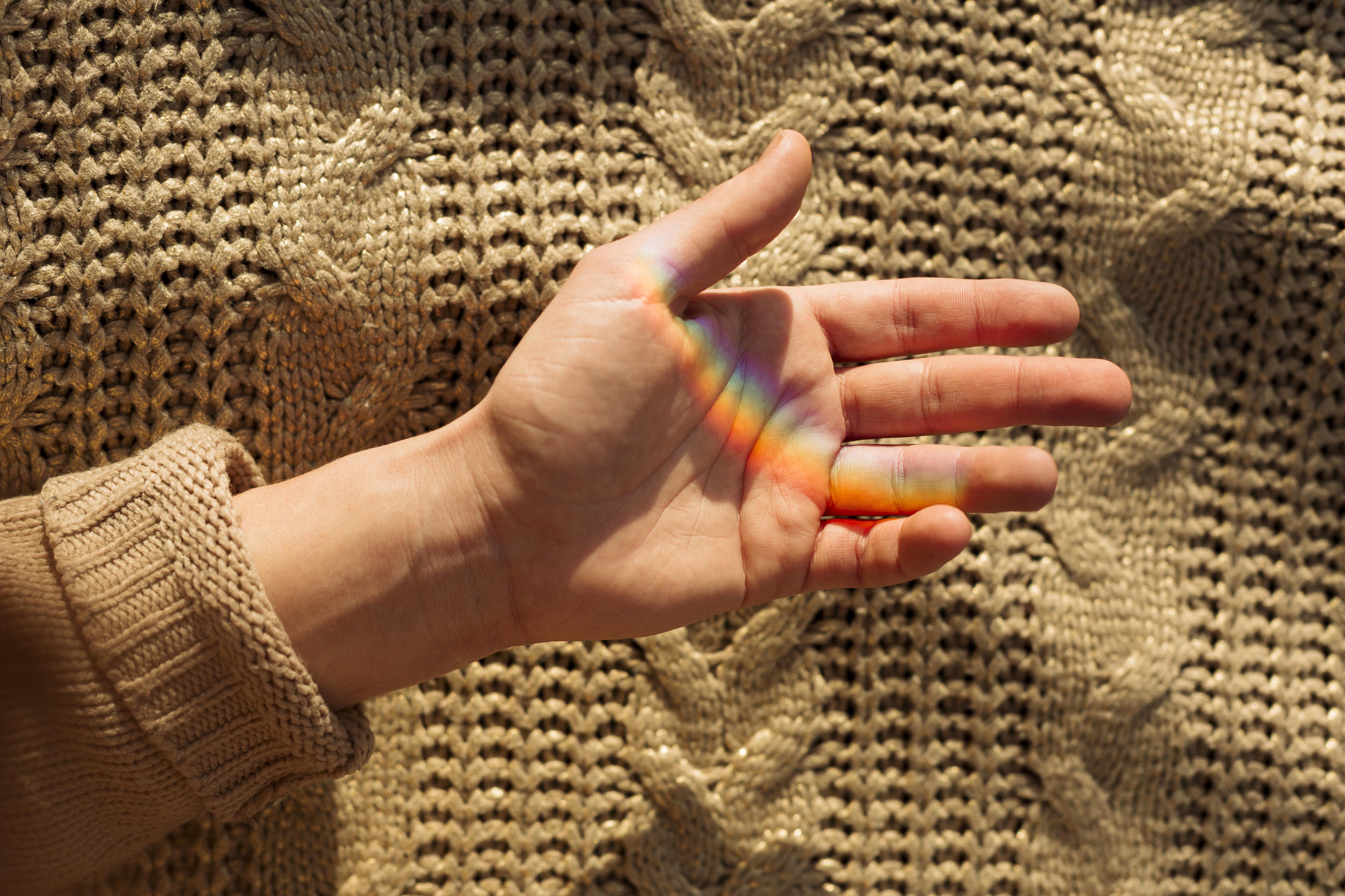 Hand mit Regenbogenlicht auf offener Handfläche vor einem beigefarbenen Strickpullover.