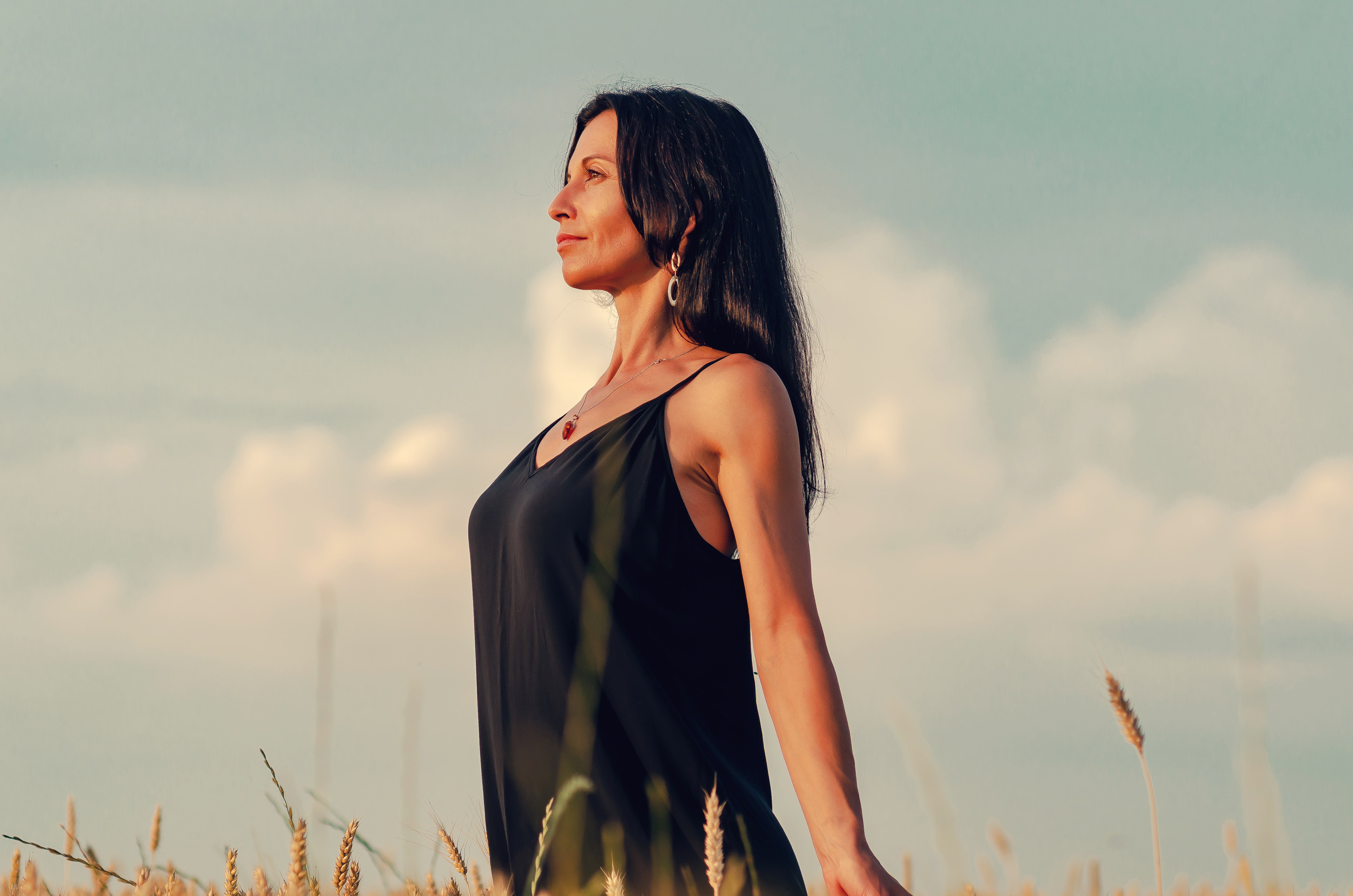 Frau mit schwarzen Haaren in schwarzem ärmellosen Kleid steht in einem Weizenfeld bei wolkigem Himmel.