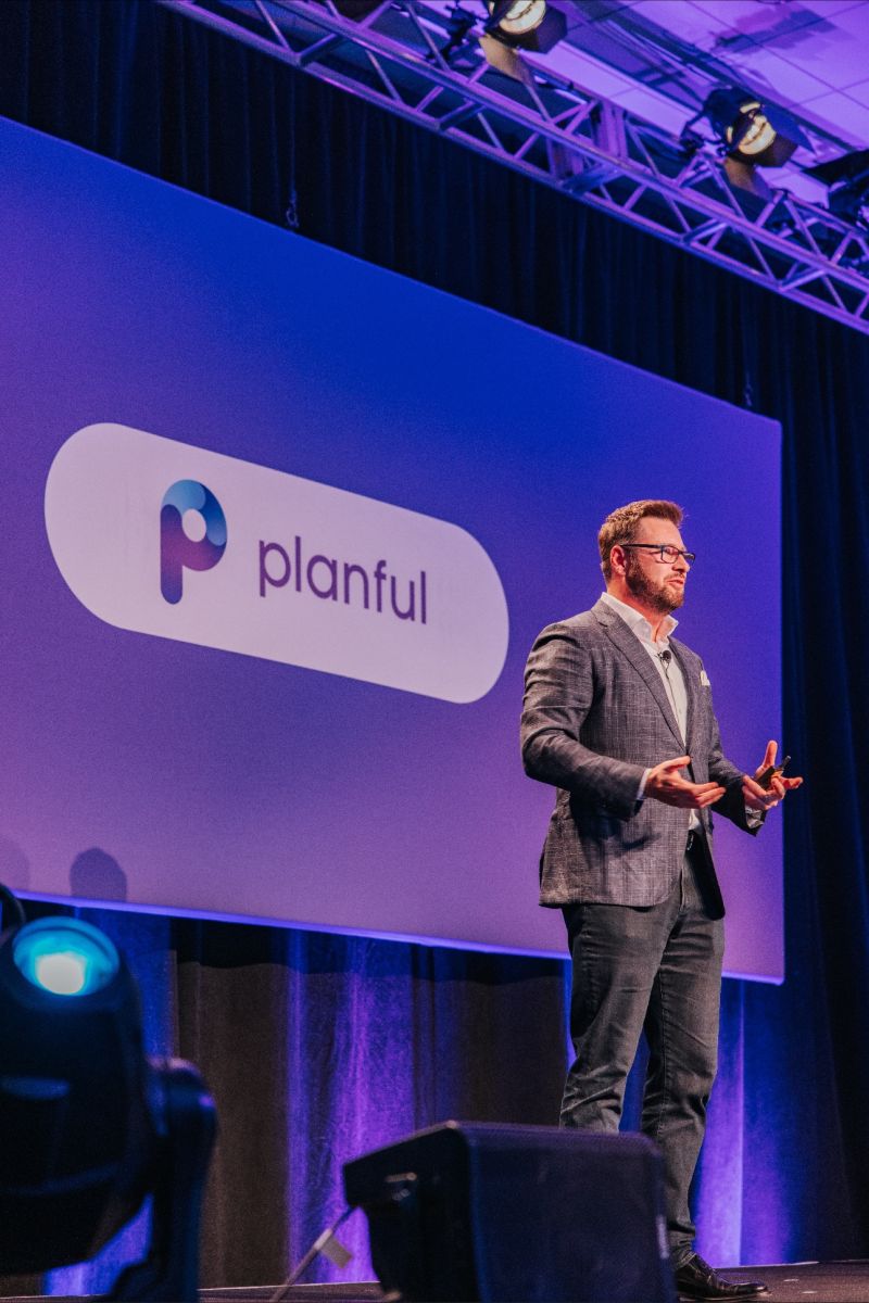 Man in a checked blazer and glasses speaking on stage with a large screen behind him displaying the Planful logo.