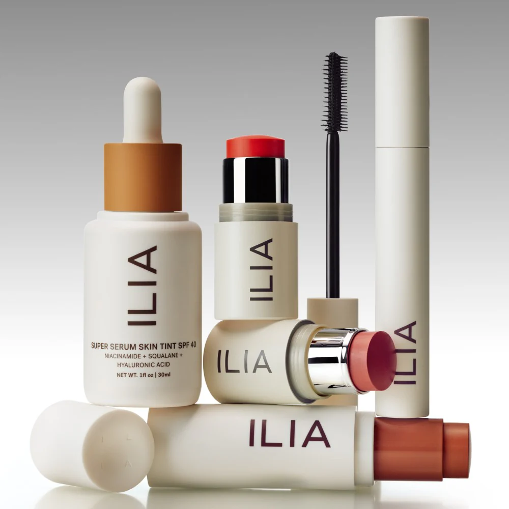 Collection of ILIA brand skincare and makeup products including serum, blush sticks, mascara, and lip balm in minimalistic beige packaging.