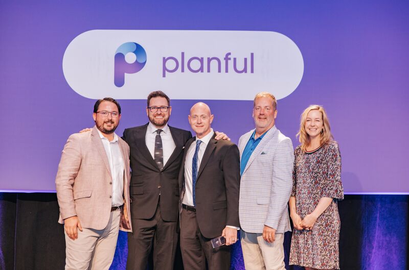 Five professionally dressed people standing together and smiling in front of a purple backdrop displaying the Planful logo.