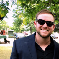 Smiling man wearing sunglasses and a dark blazer outdoors with trees and parked cars in the background.