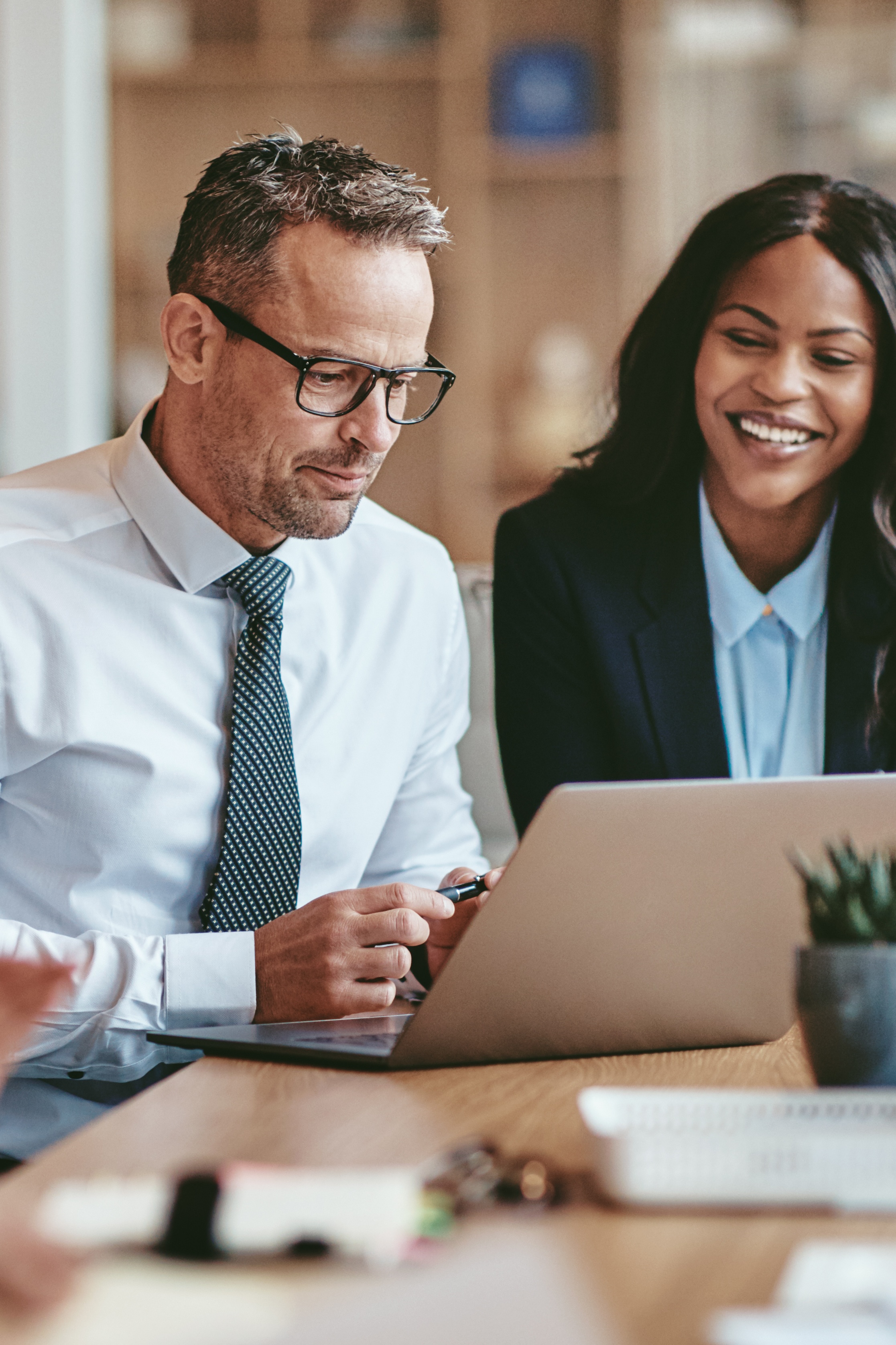 Two business professionals, a man and a woman, looking and smiling at a laptop screen together in an office setting.
