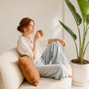 Red-haired woman in white shirt and jeans sitting on couch, drinking from brown mug, next to large houseplant.