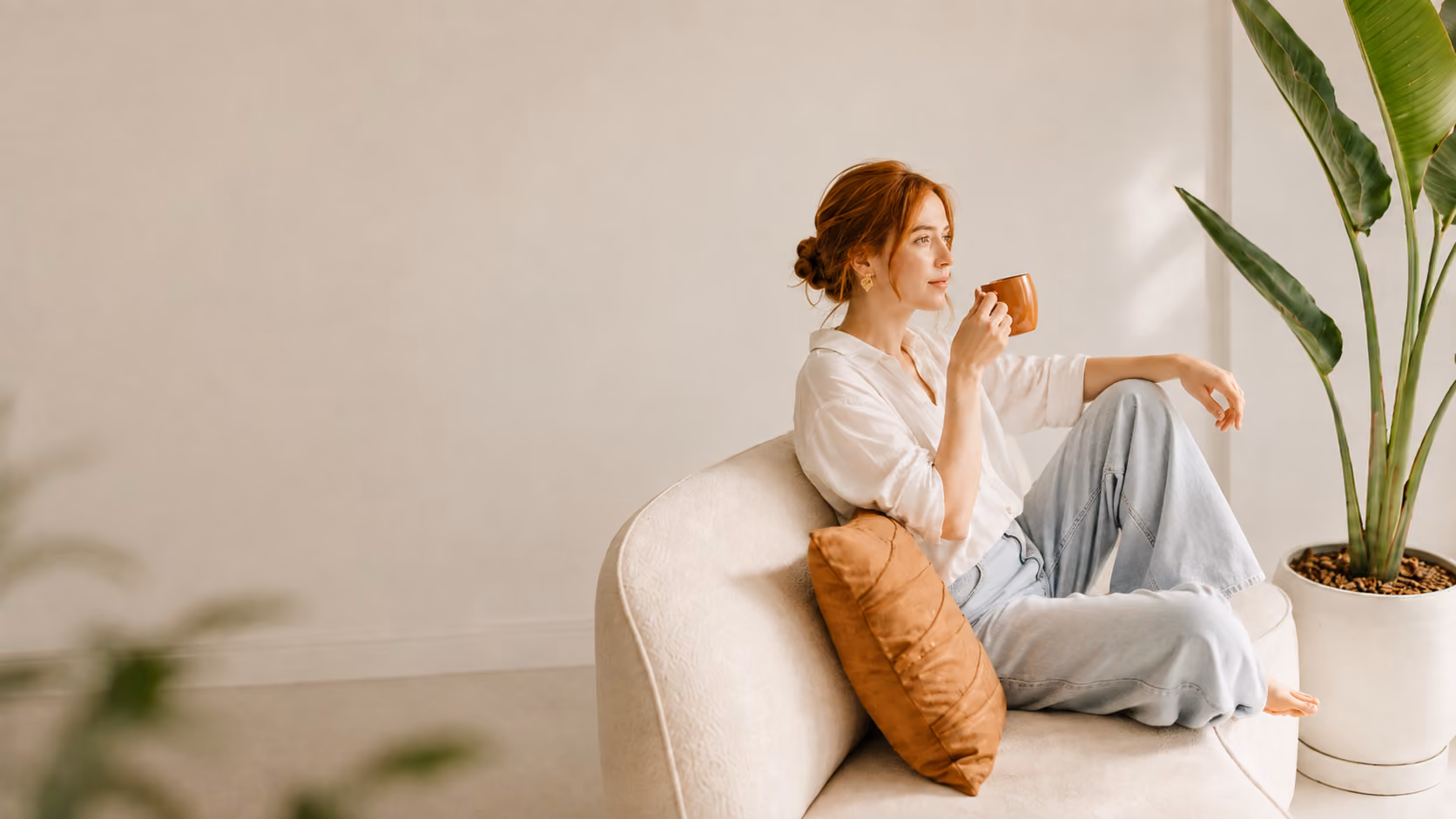 Red-haired woman in white shirt and jeans sitting on couch, drinking from brown mug, next to large houseplant.