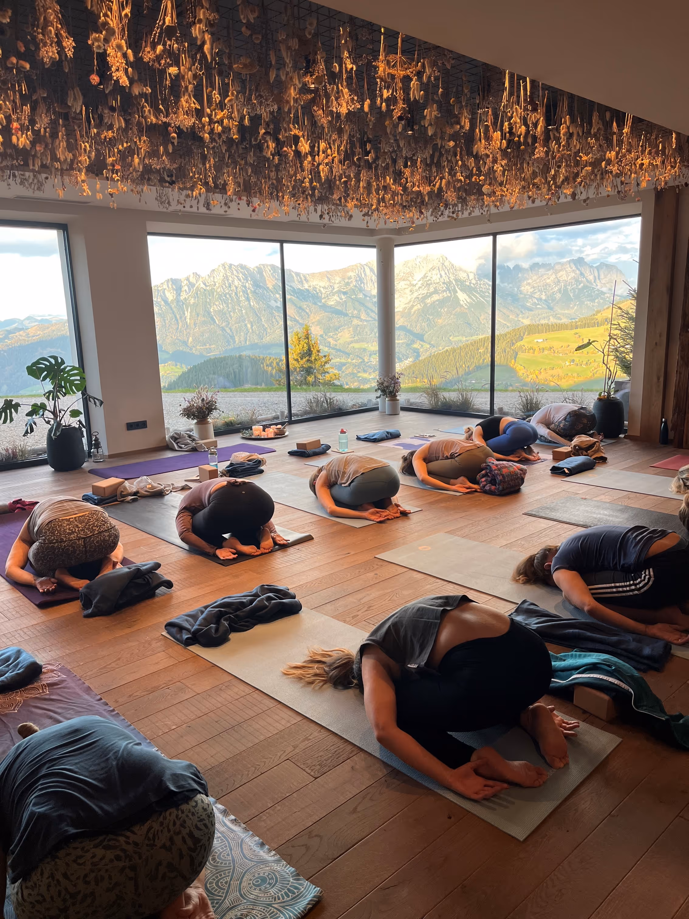 Menschen praktizieren Yoga in Vorwärtsbeuge in einem hellen Raum mit großen Fenstern und Bergblick.