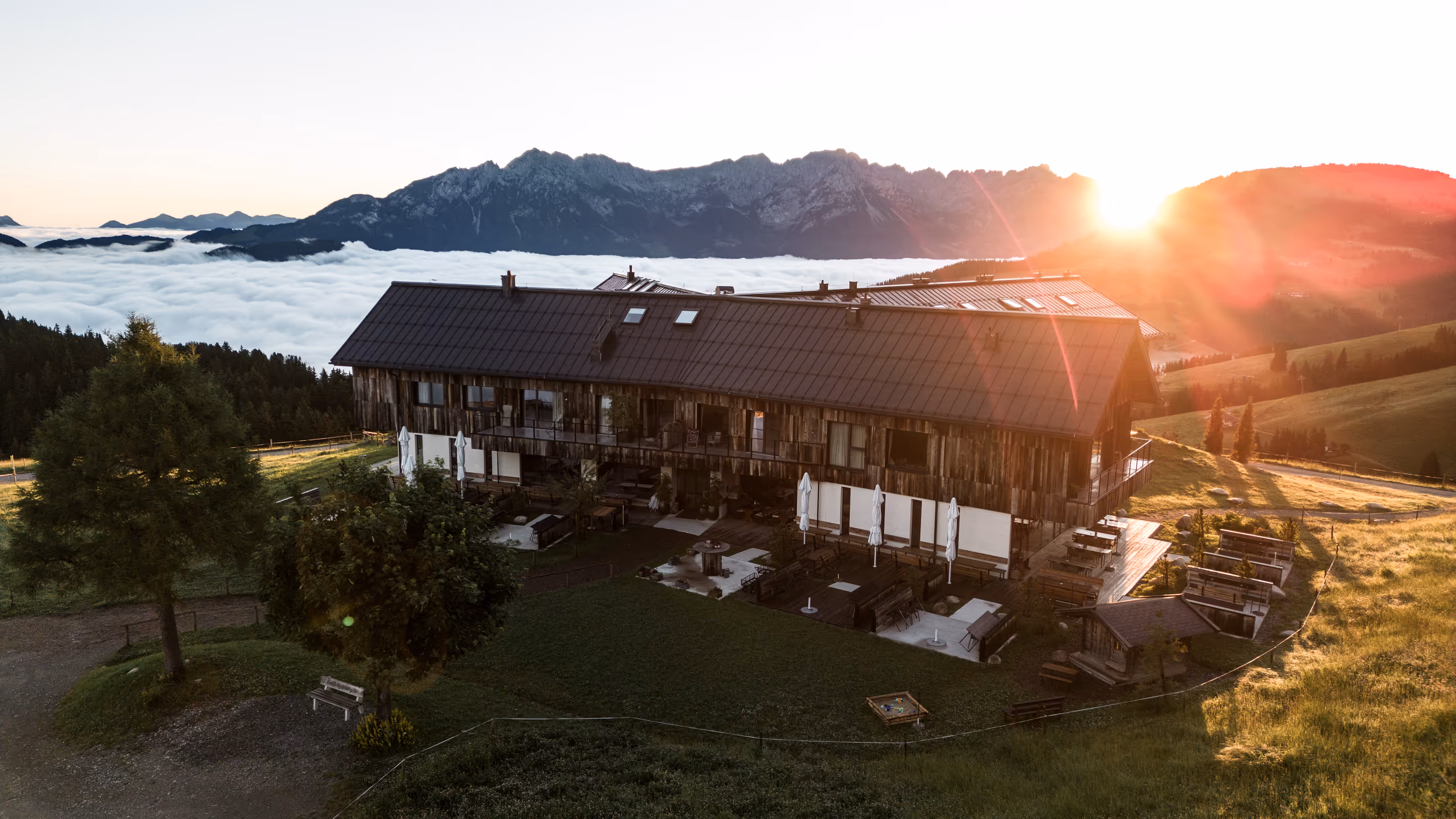 Almhütte bei Sonnenuntergang mit grünen Wiesen, zwei Bäumen und Bergen im Hintergrund über einer Nebeldecke.