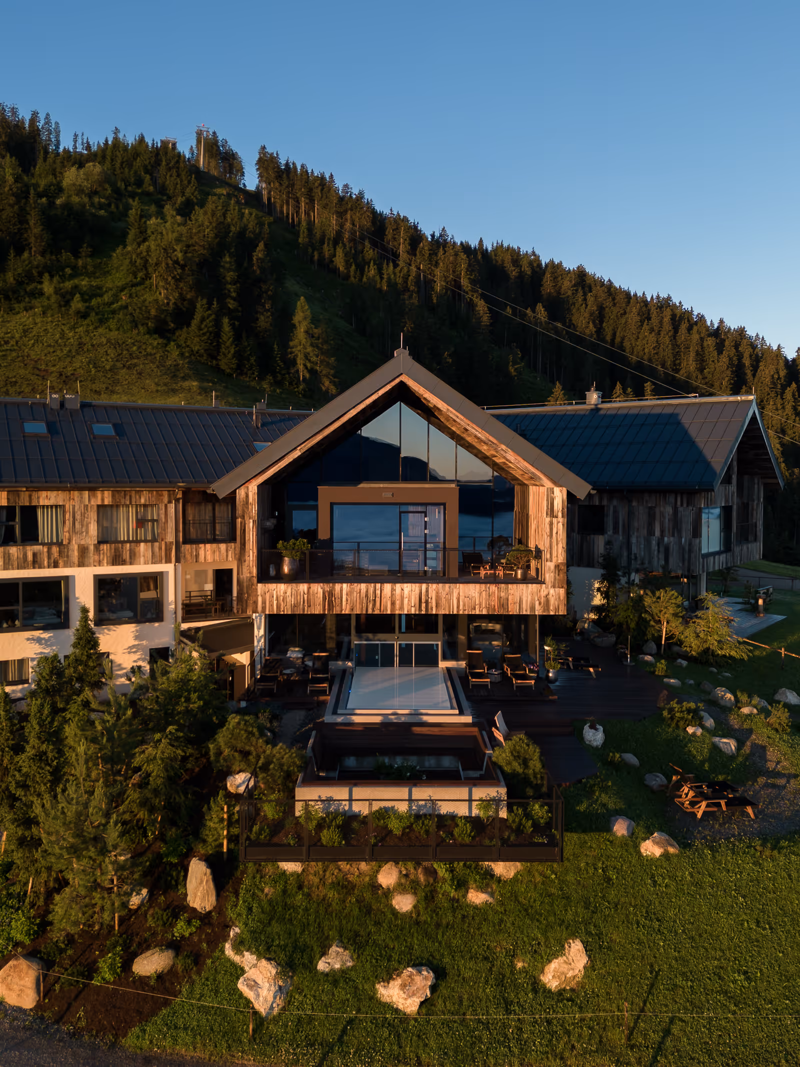 Modernes Berghotel mit Holzfassade, Terrasse und Blick auf bewaldeten Hügel bei klarem Himmel.