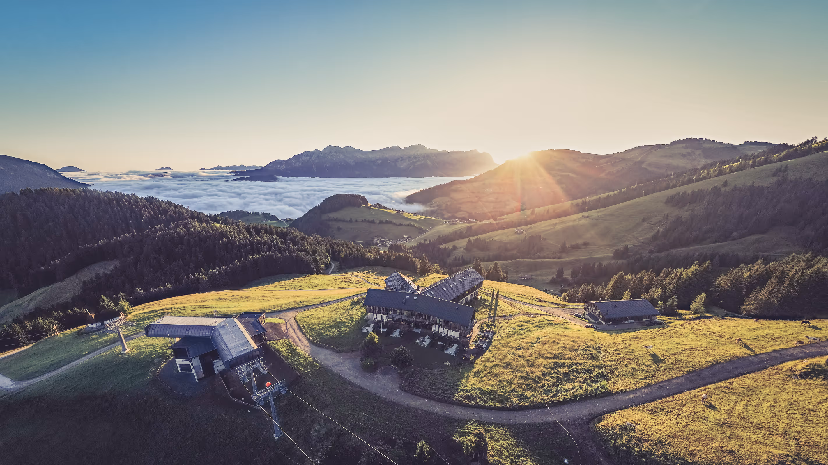 Sonnenaufgang über einem Bergresort mit grünen Wiesen, Wäldern und Nebel in einem Tal mit Bergen im Hintergrund.