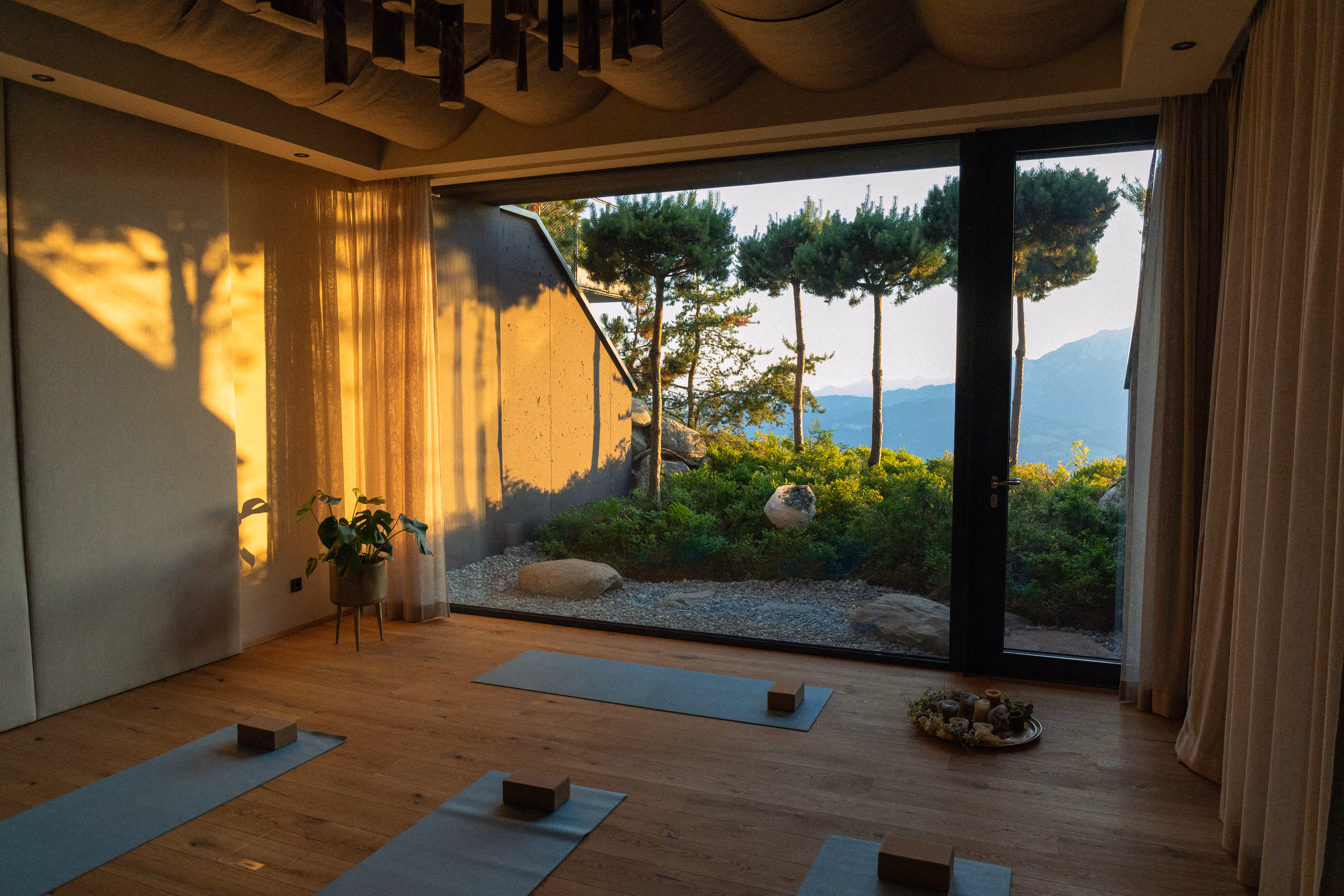 Ruhiger Yoga-Raum mit Holzfußboden, Yoga-Matten und Blöcken vor einer großen Glaswand mit Blick auf Bäume und Berge im Sonnenuntergang.