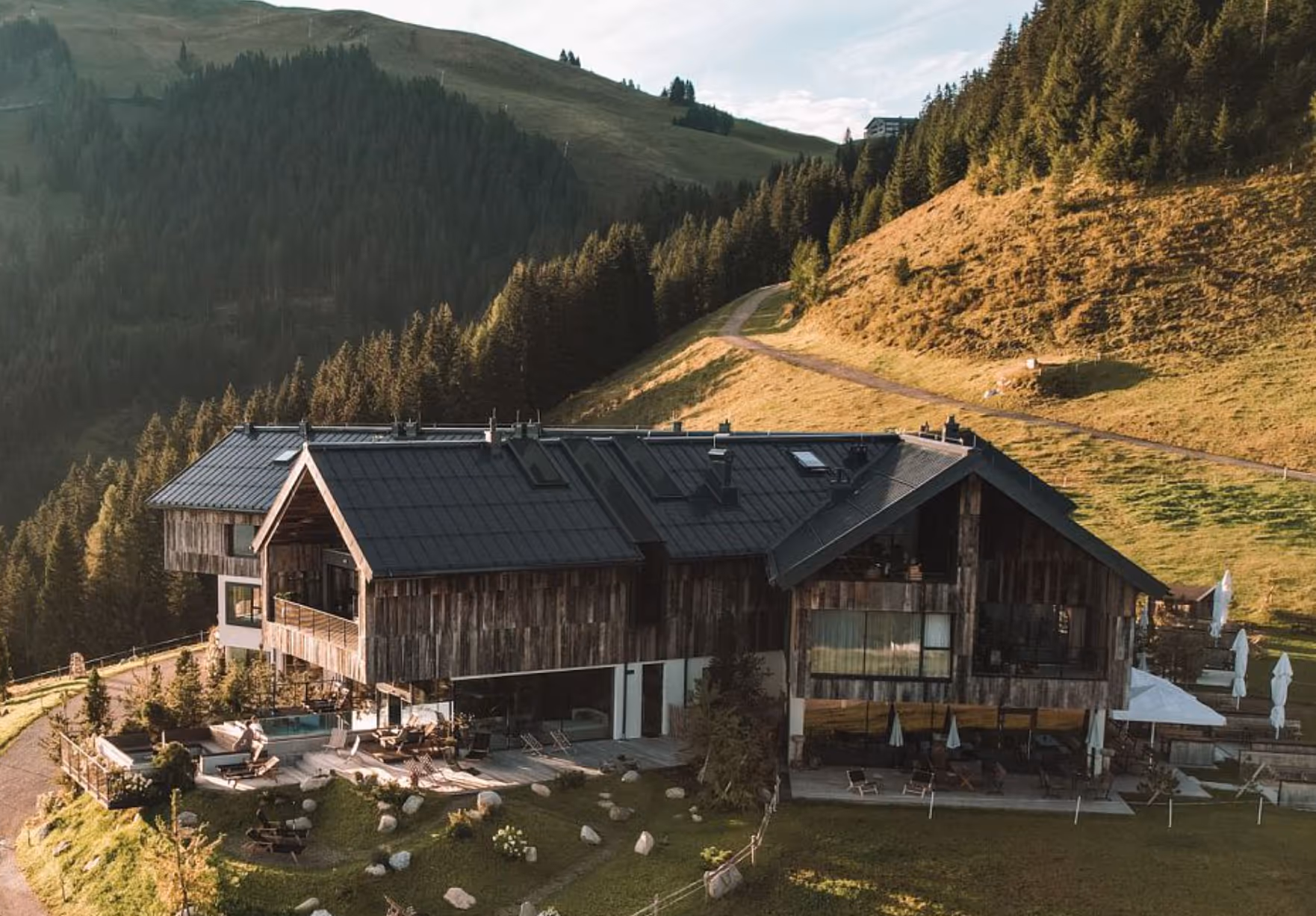 Großes Holzhotel mit Terrasse und Liegestühlen in einer bergigen Waldlandschaft bei Sonnenlicht.