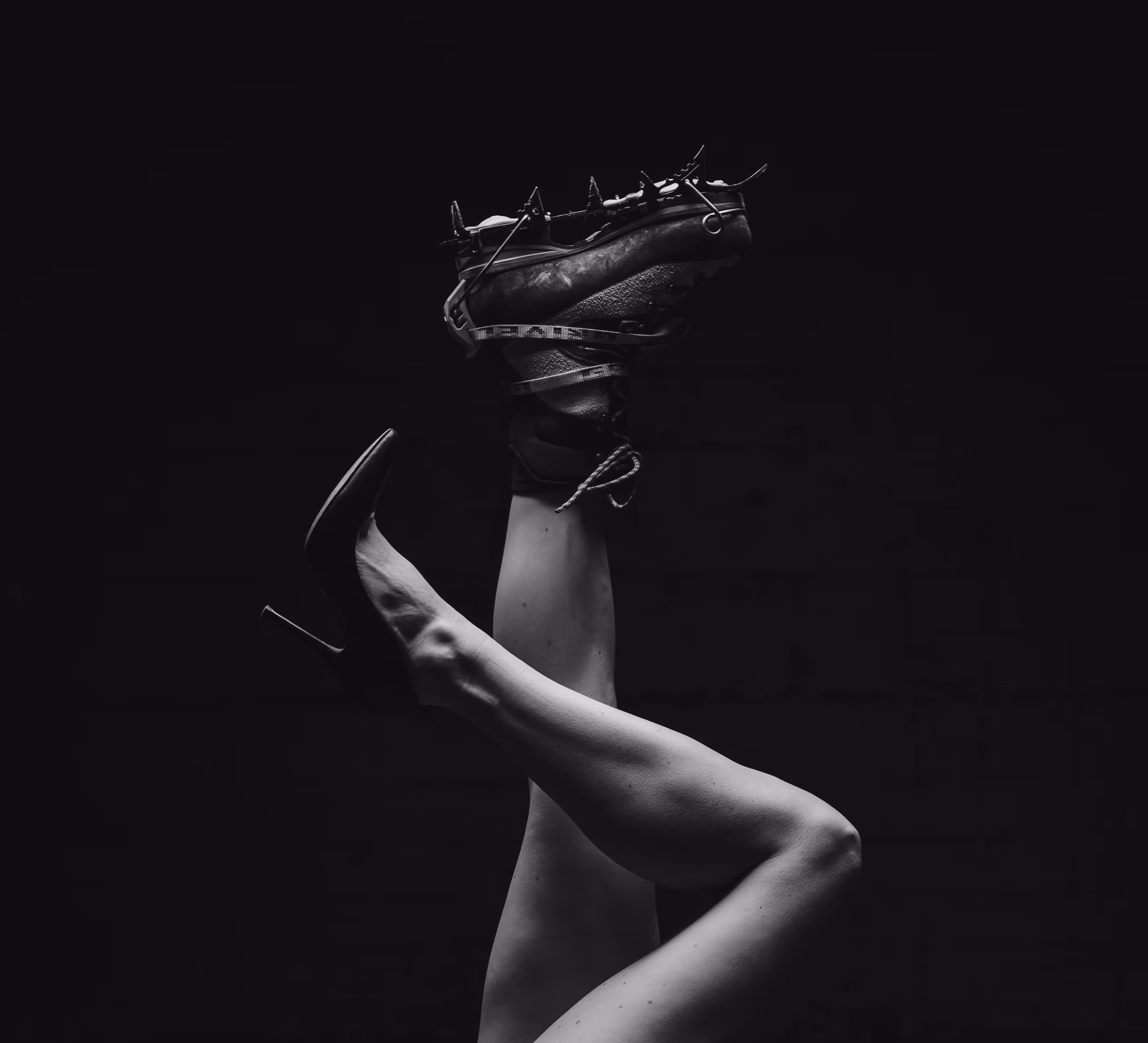 Schwarz-weiß-Foto von gekreuzten Beinen, ein Bein trägt einen High Heel, das andere einen Spike-Sportschuh.