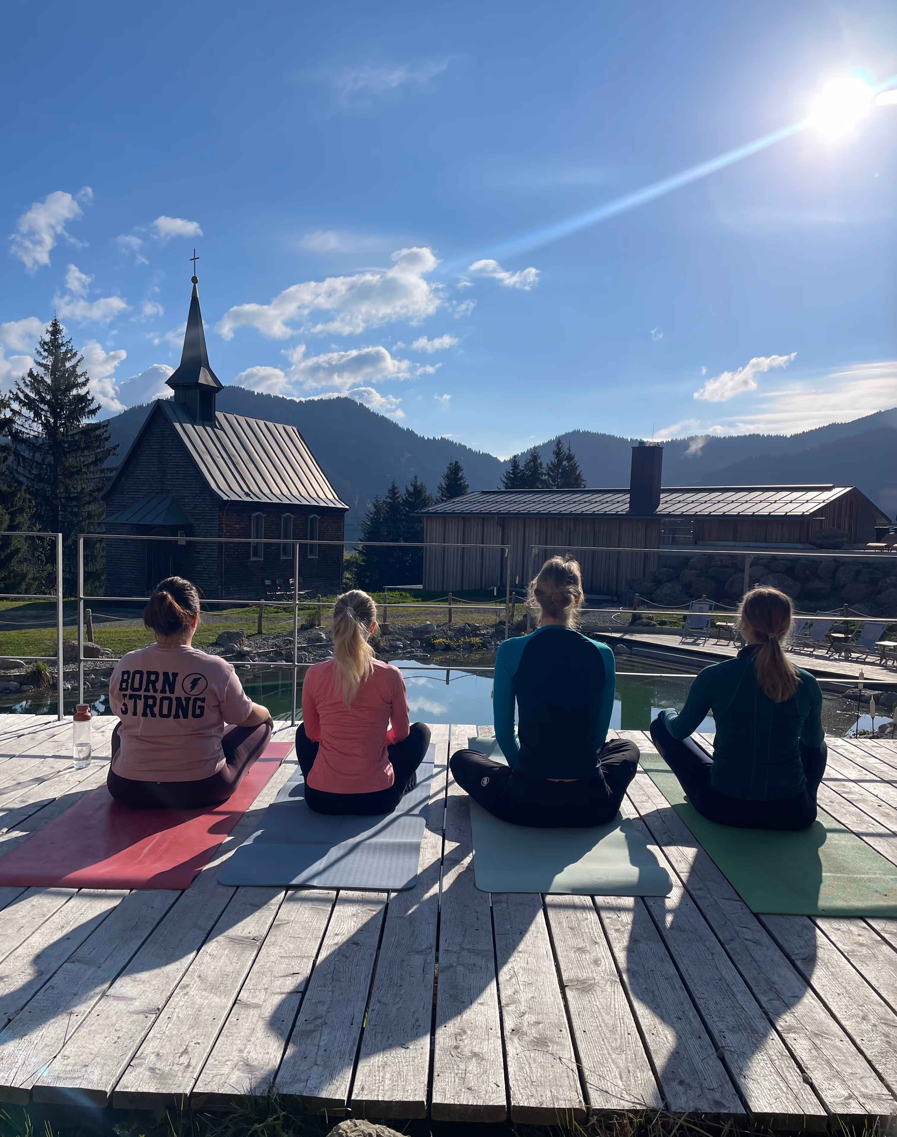 Vier Frauen sitzen auf Yogamatten auf einer Holzplattform, vor einer Berglandschaft mit Kirche und Gebäuden bei sonnigem Himmel.