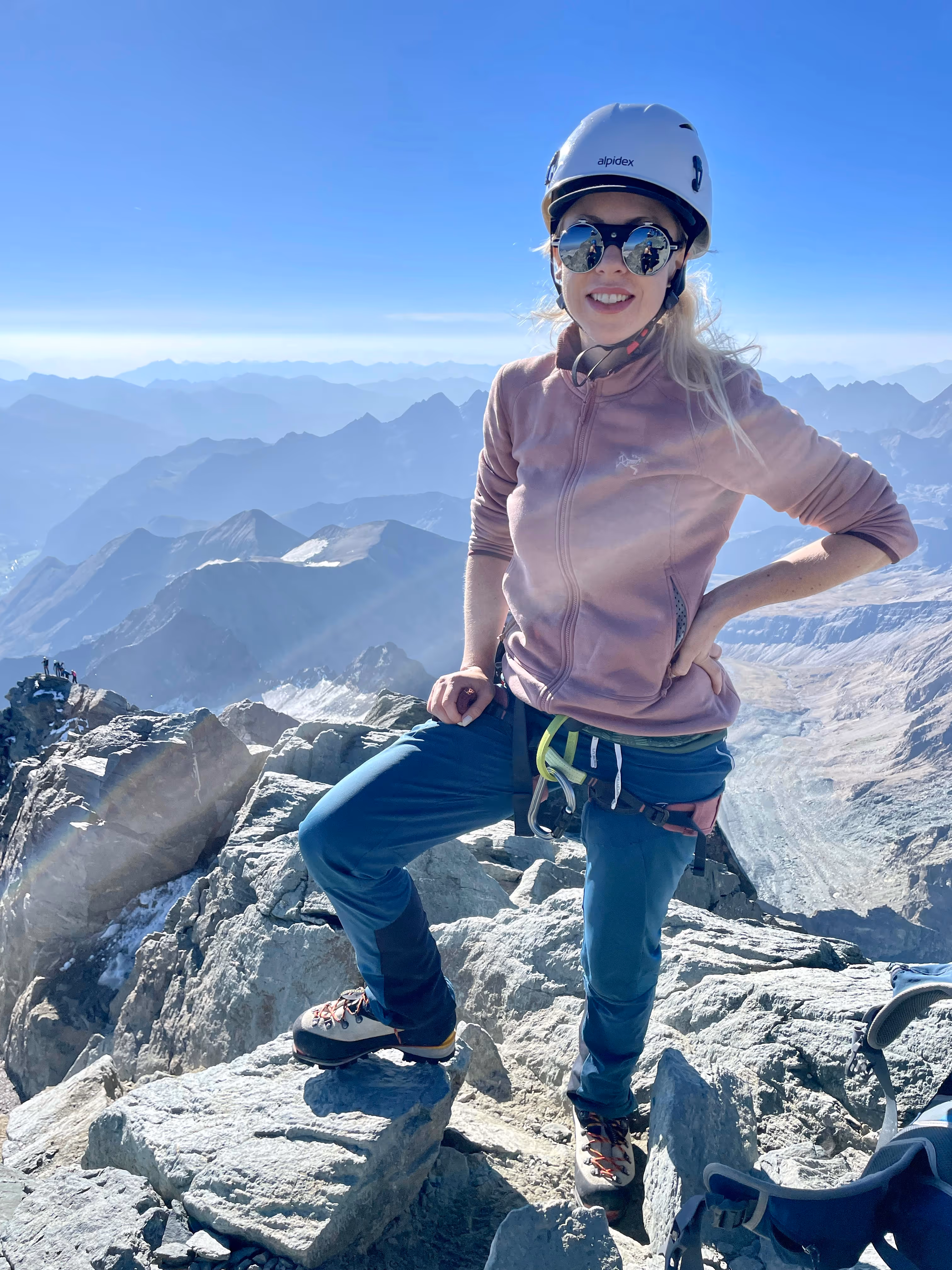 Frau mit Helm und Sonnenbrille steht lächelnd auf felsigem Berggipfel mit Bergpanorama im Hintergrund.
