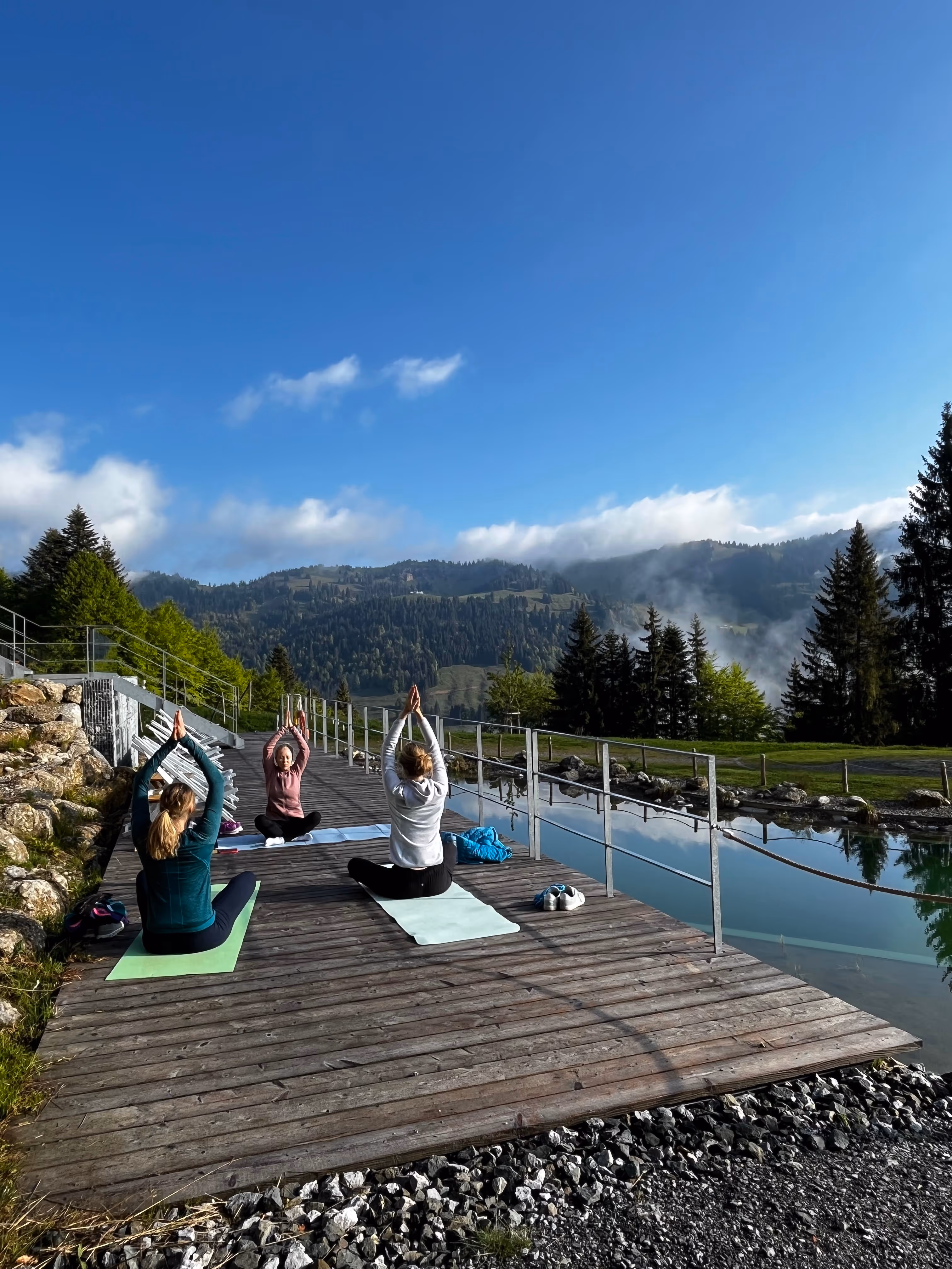 Drei Personen praktizieren Yoga auf einer Holzplattform neben einem Teich mit Blick auf bewaldete Berge unter blauem Himmel.