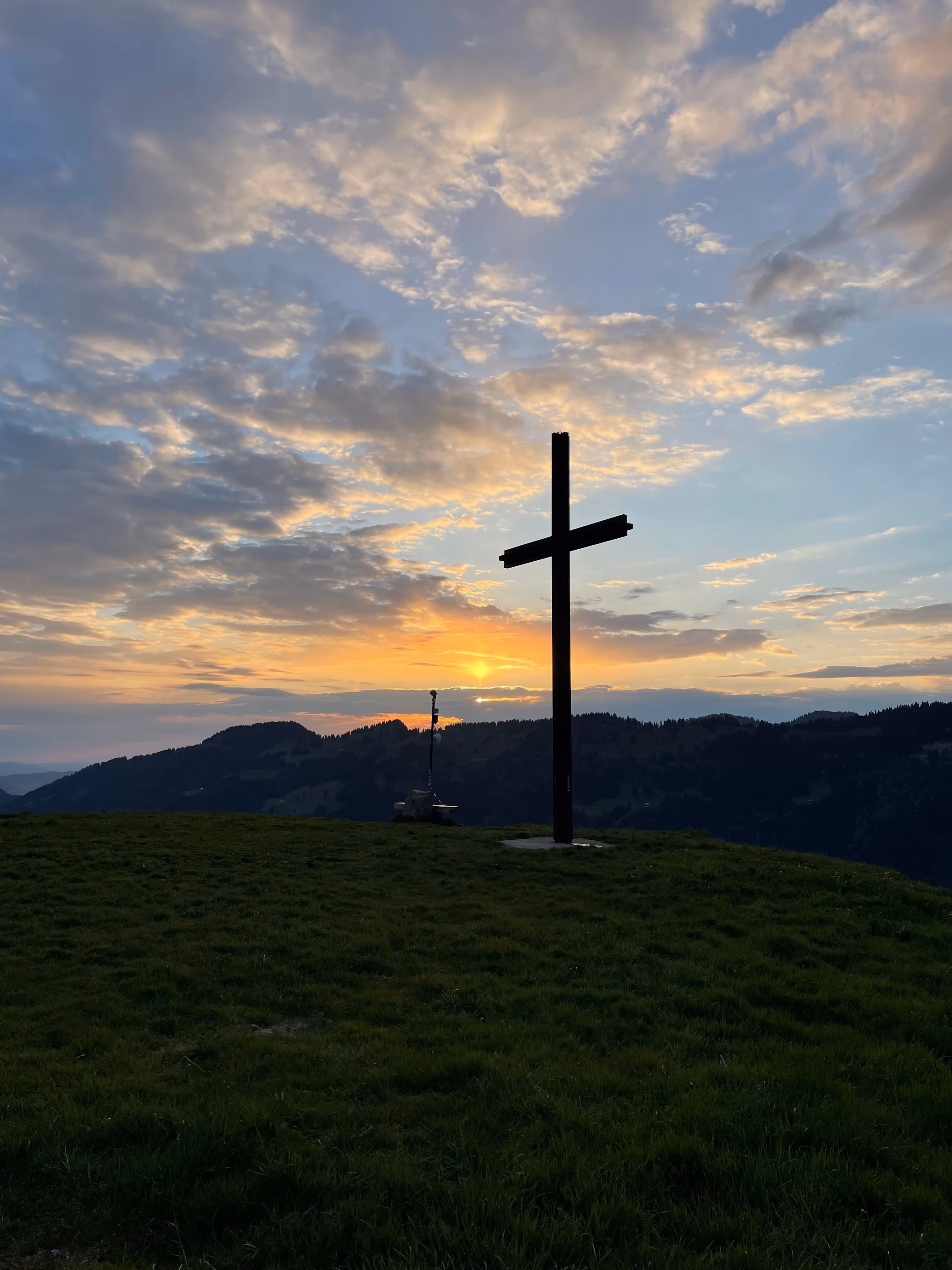 Holzkreuz auf einem Hügel bei Sonnenuntergang mit bewölktem Himmel und Bergen im Hintergrund.