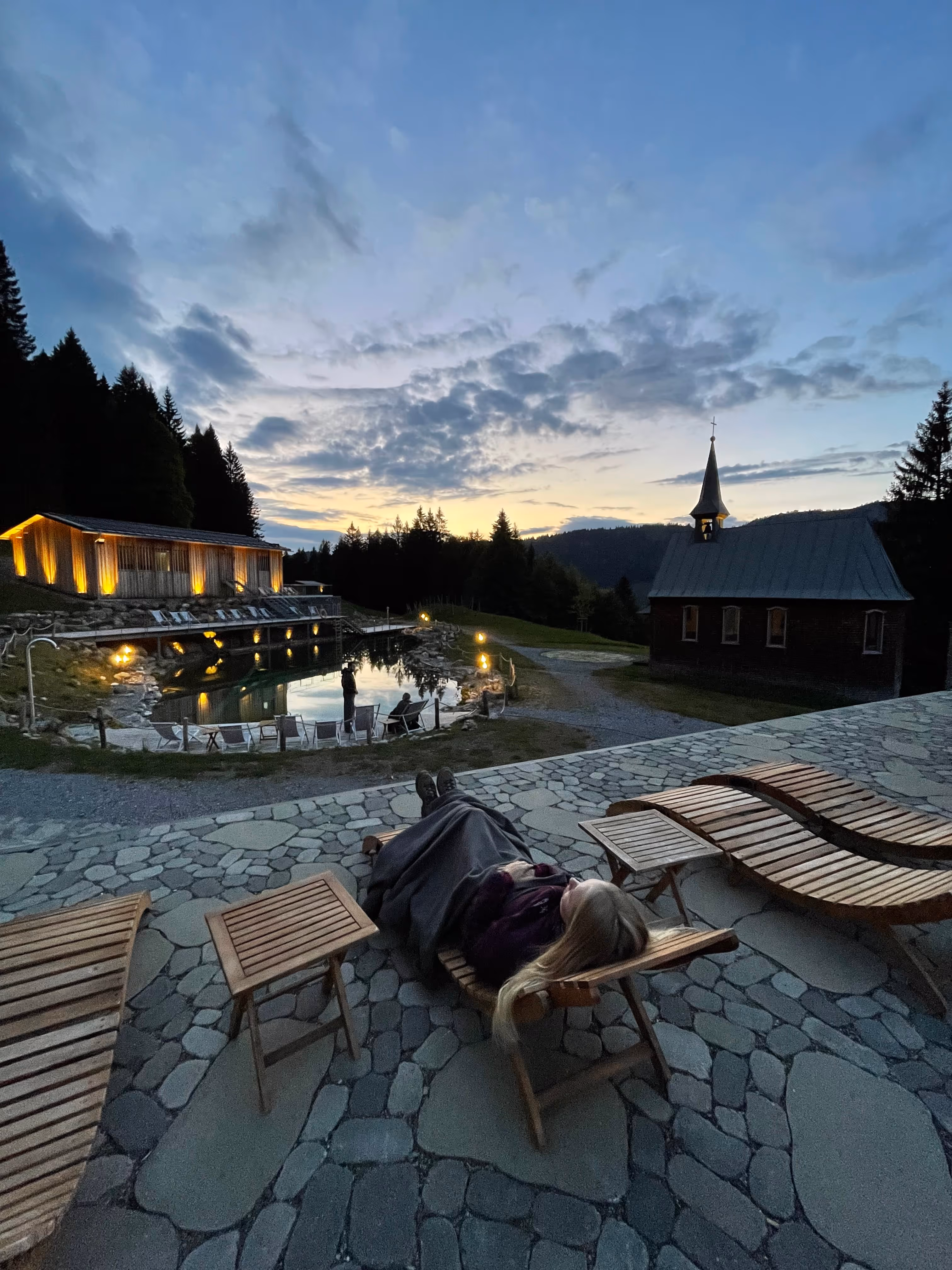 Person liegt mit einer Decke auf einer Holzliege auf gepflastertem Boden bei Sonnenuntergang, mit beleuchtetem Gebäude, Teich und Kirchgebäude im Hintergrund.