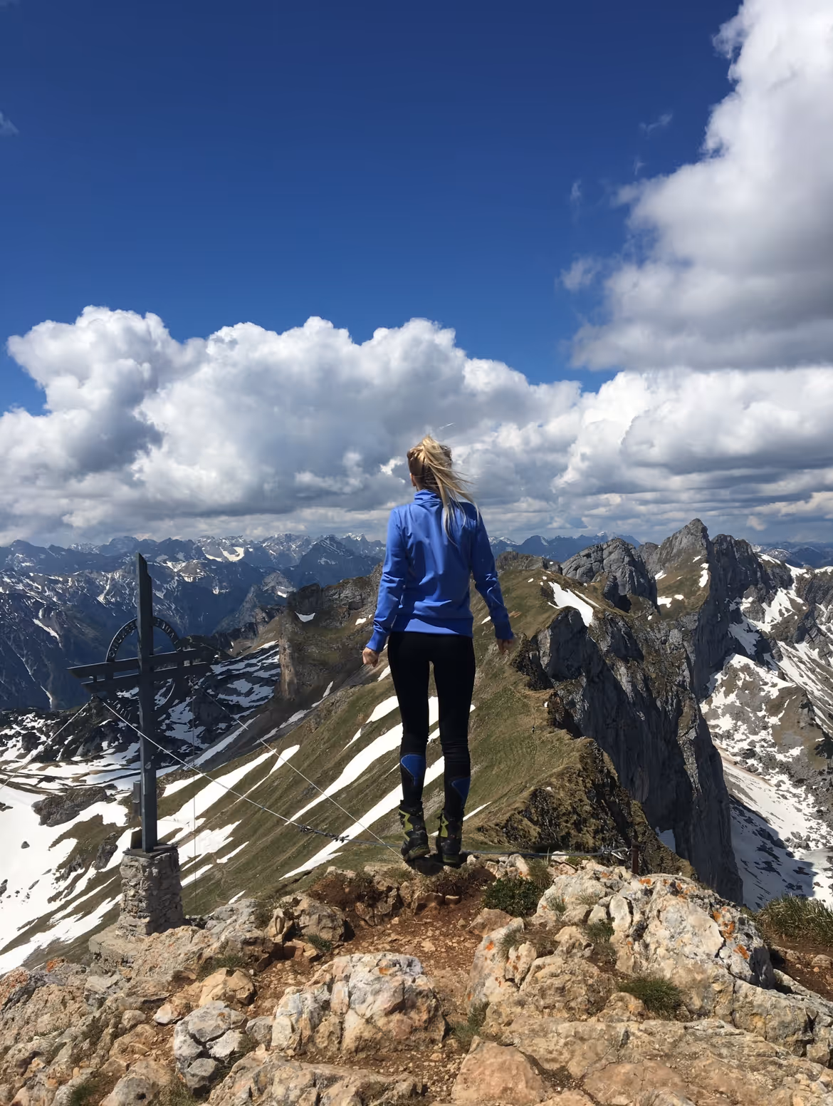 Eine Person in blauer Jacke steht auf einem felsigen Gipfel mit Blick auf schneebedeckte Bergkämme unter blauem Himmel mit Wolken.