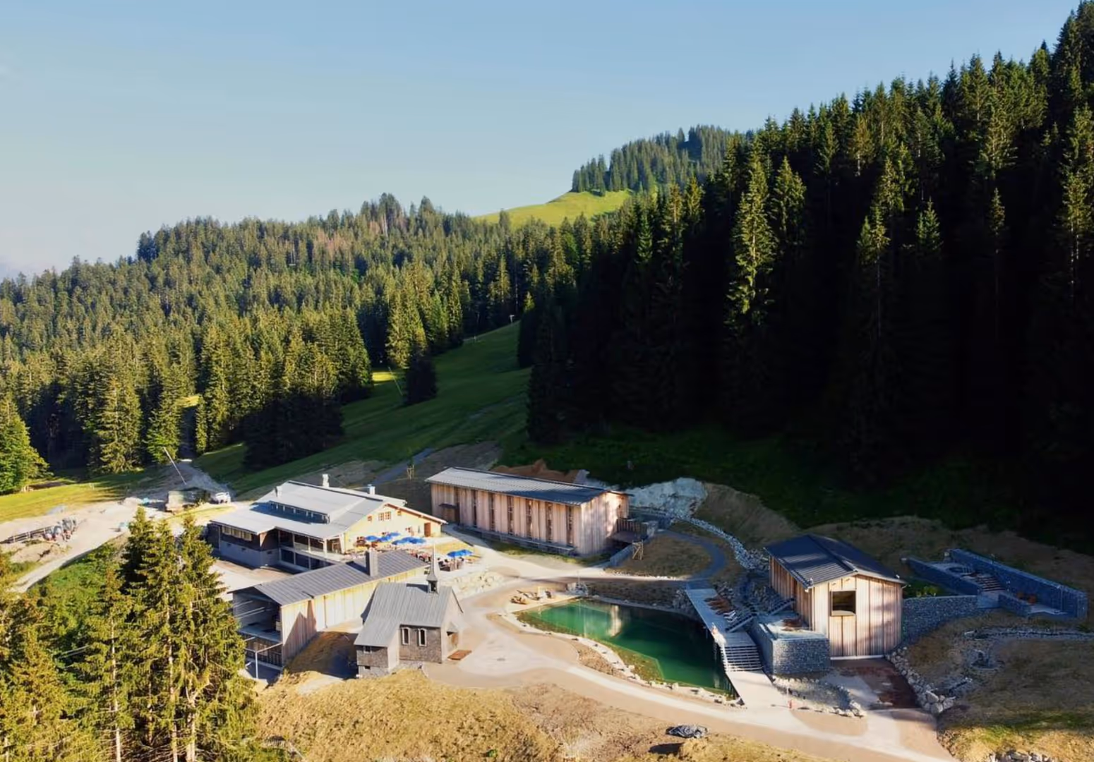 Bergresort mit mehreren Holzhäusern neben einem natürlichen Badeteich, umgeben von einer Nadelwaldlandschaft bei klarem Himmel.