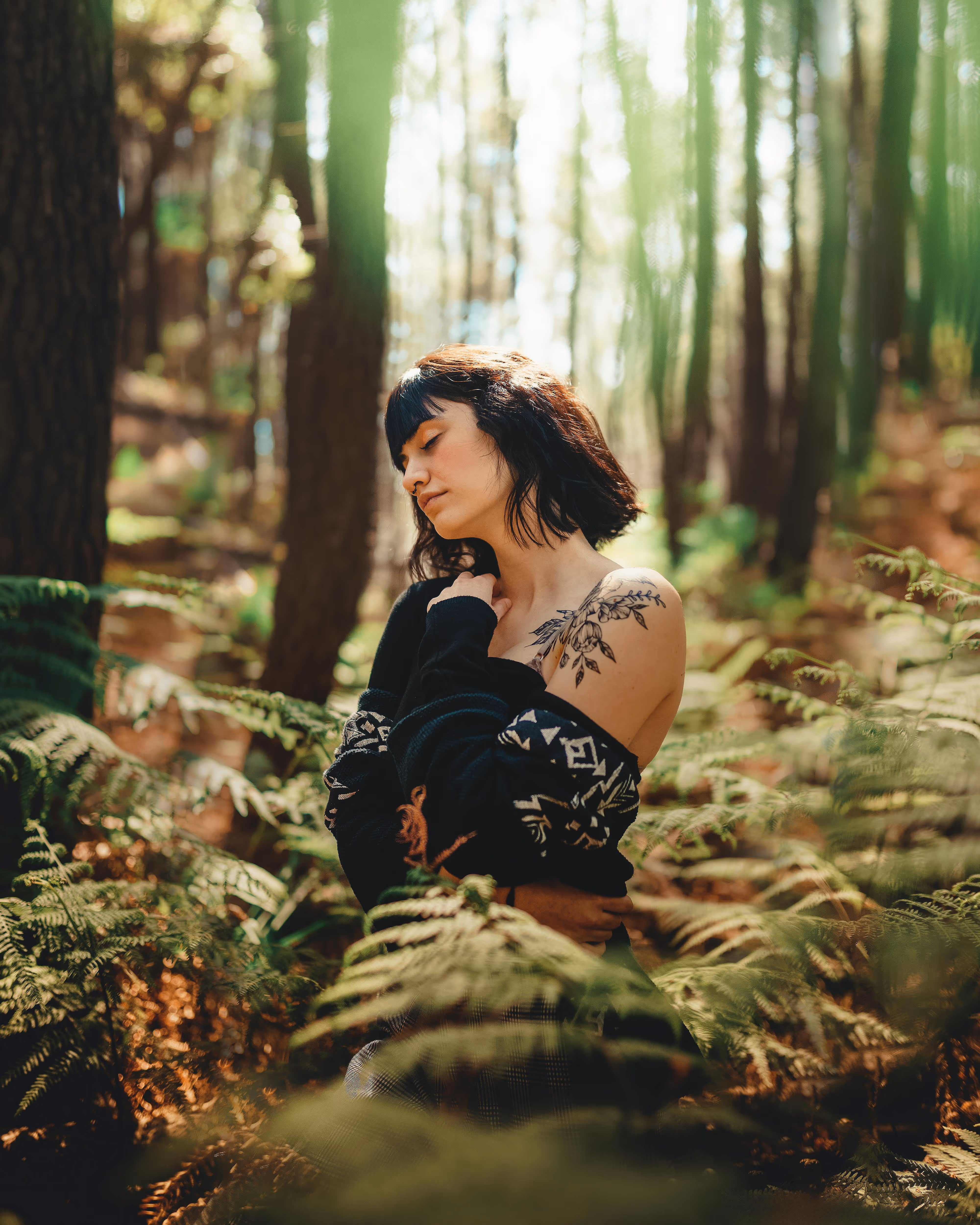 Frau mit Tattoo auf der Schulter sitzt entspannt in einem sonnendurchfluteten Wald zwischen Farnen.