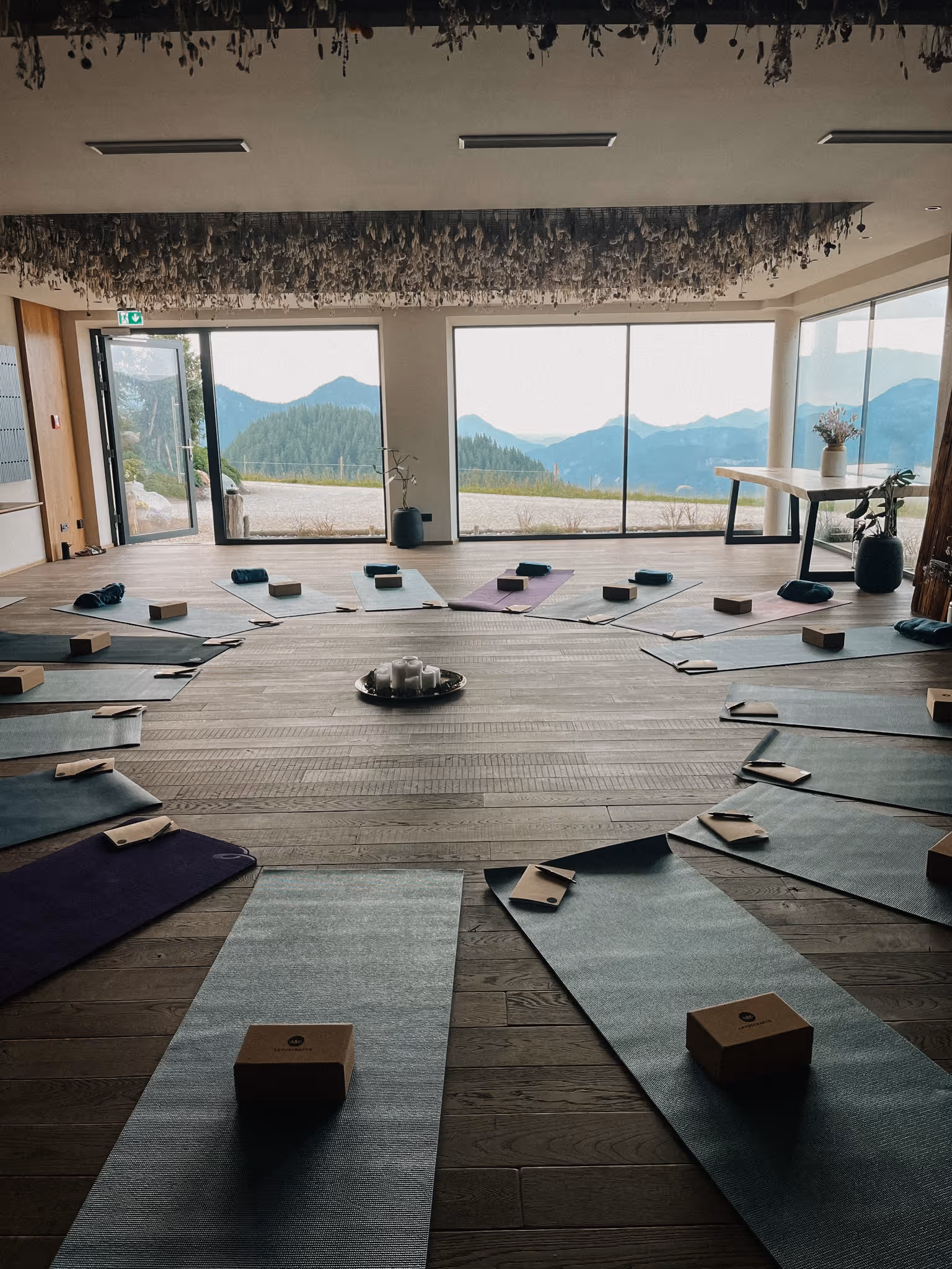 Yoga-Studio mit kreisförmig angeordneten Matten, Blöcken und Notizbüchern vor großen Fenstern mit Bergblick.