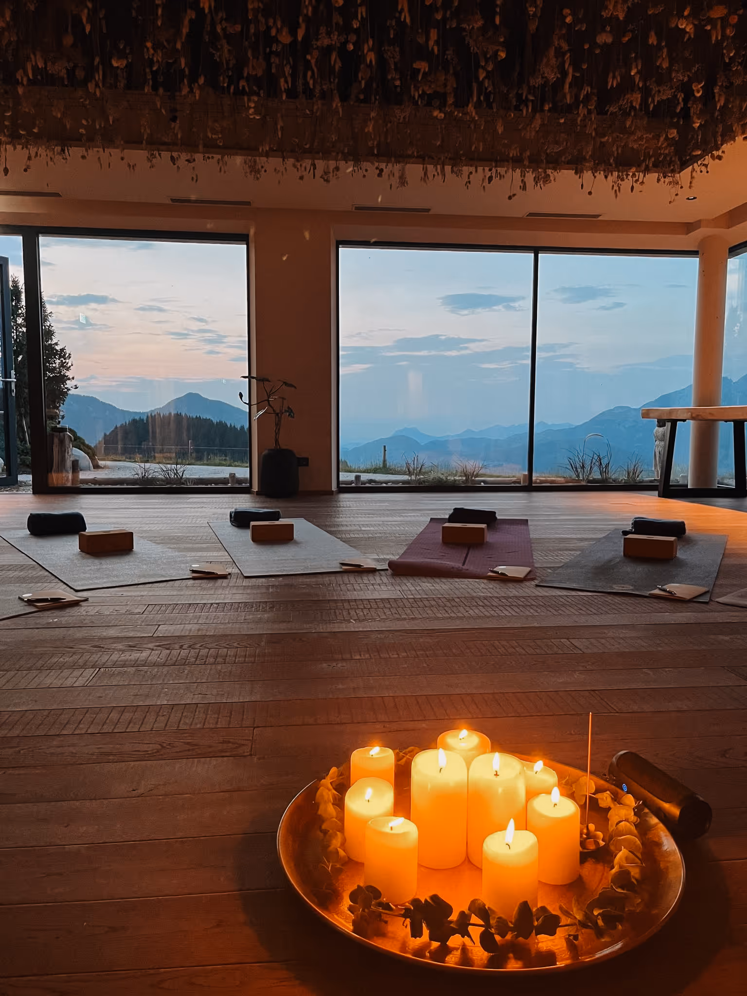 Sanfter Yoga-Raum mit brennenden Kerzen in einer Schale, Yogamatten und Klötzen vor großen Fenstern mit Bergblick bei Sonnenuntergang.