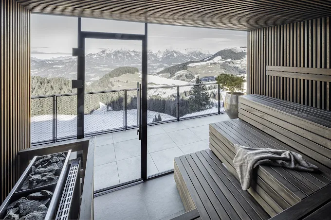 Moderne Sauna mit Holzverkleidung, Stufen mit Handtuch und Aussicht auf schneebedeckte Berge durch eine Glaswand.