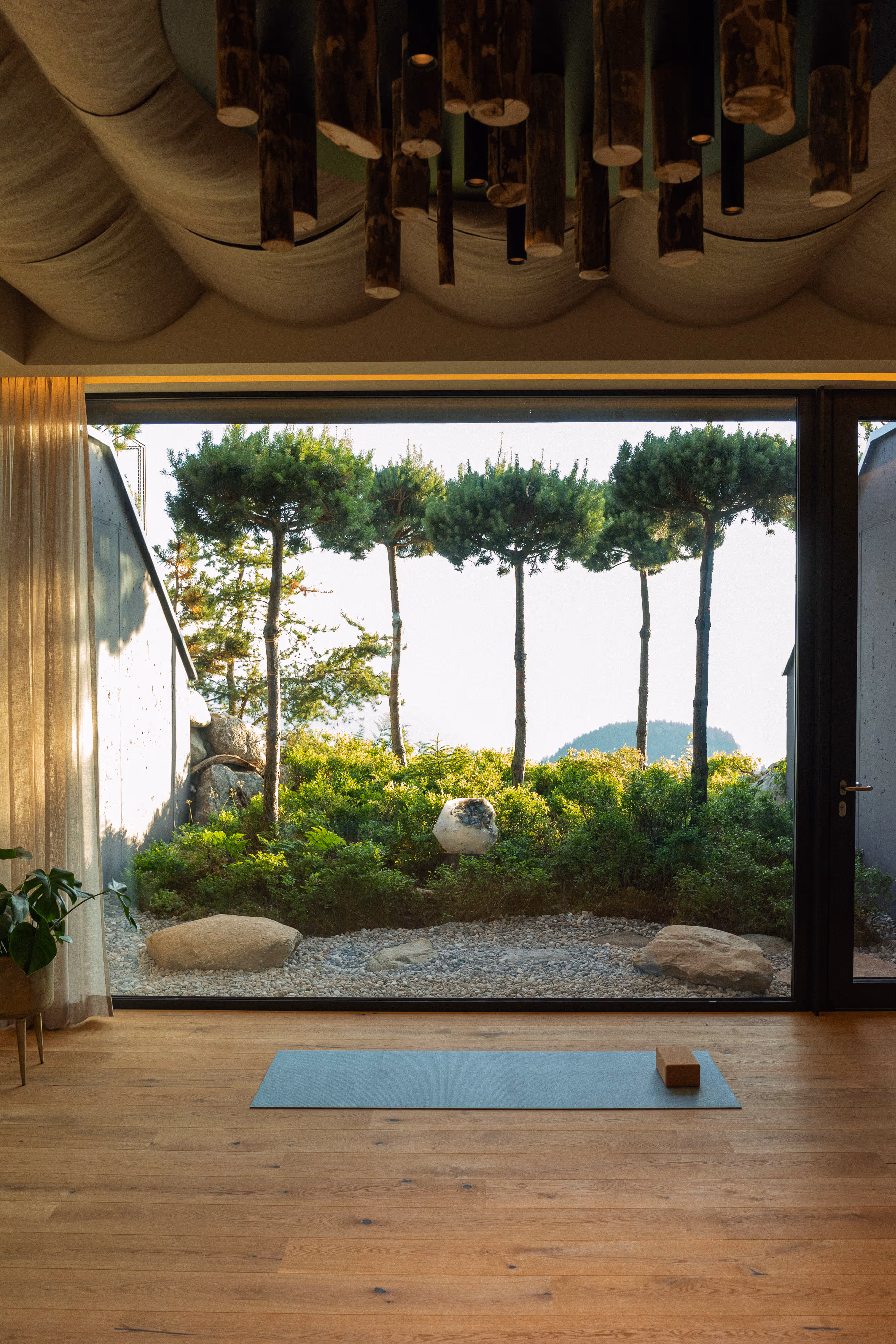 Moderner Raum mit Holzboden, Yogamatte und Block vor großem Fenster mit Blick auf Gartengrün und schlanke Bäume.