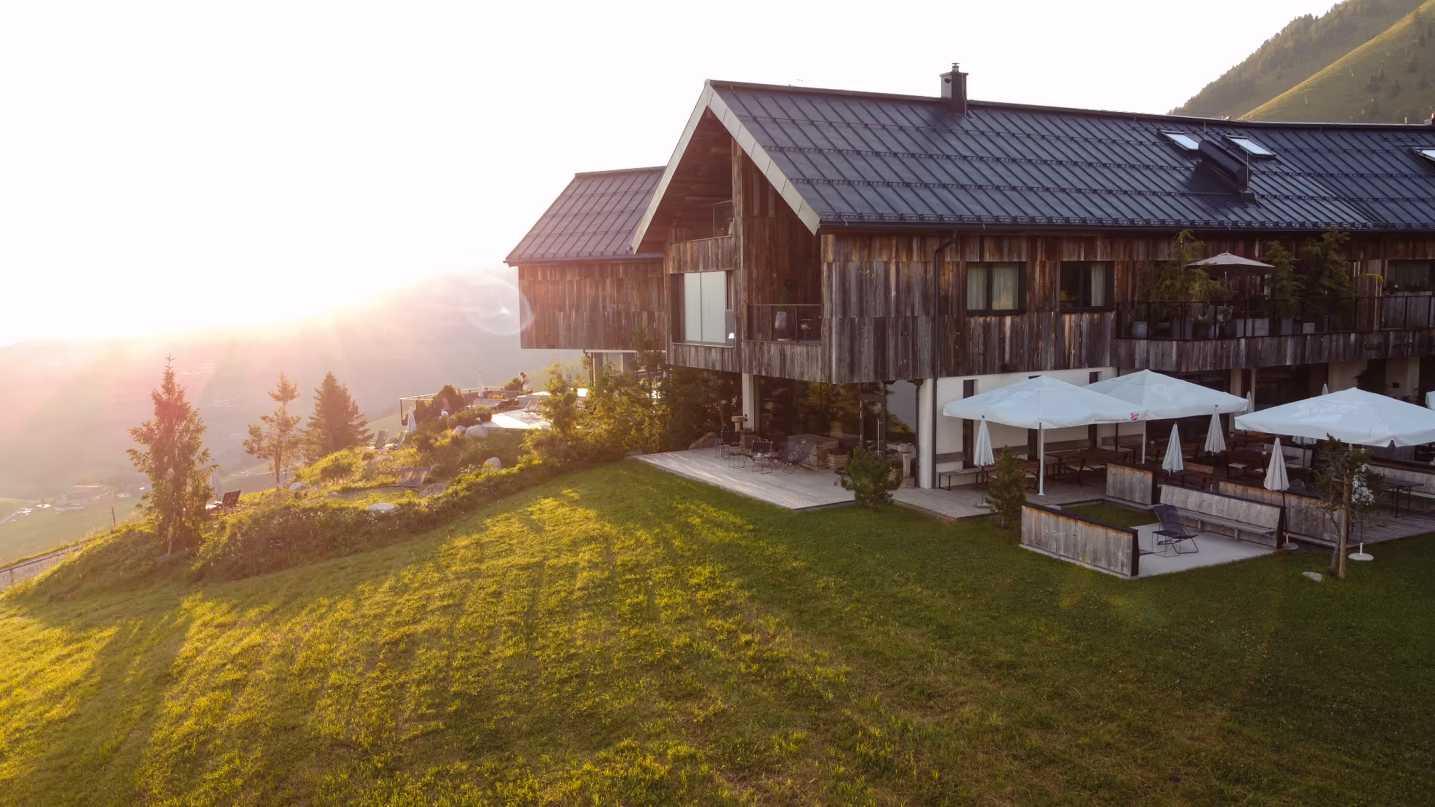 Modernes Holzhaus mit großer Terrasse und Sonnenschirmen auf einem grünen Hügel bei Sonnenuntergang in den Bergen.