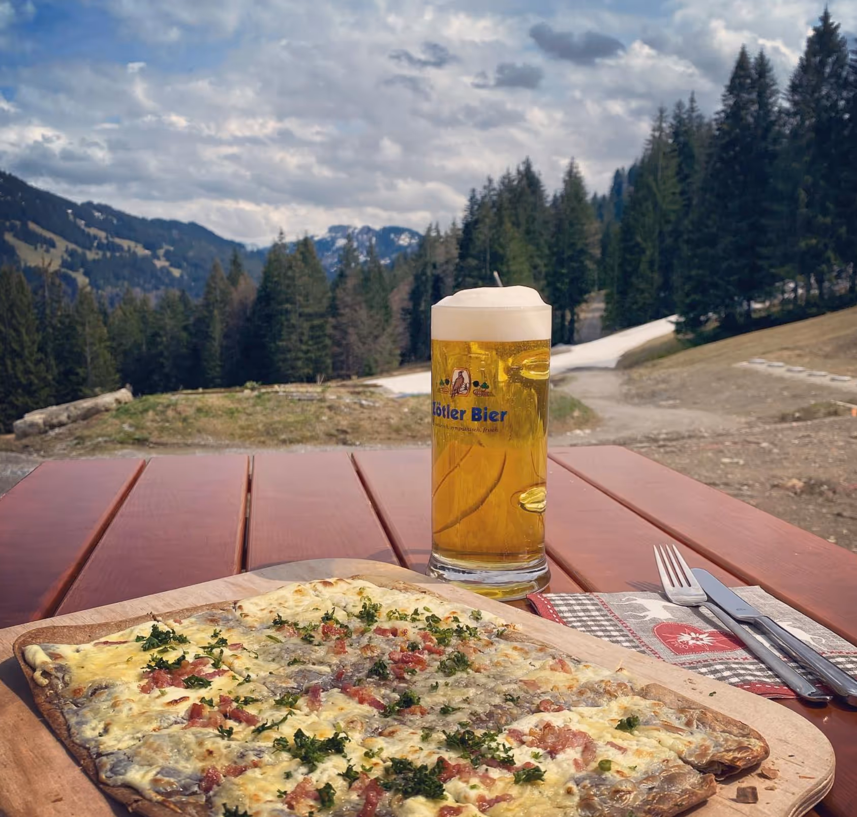 Brett mit Flammkuchen und ein Glas Lößler Bier auf einem Holztisch in Alpenlandschaft mit Nadelbäumen und Bergen im Hintergrund.