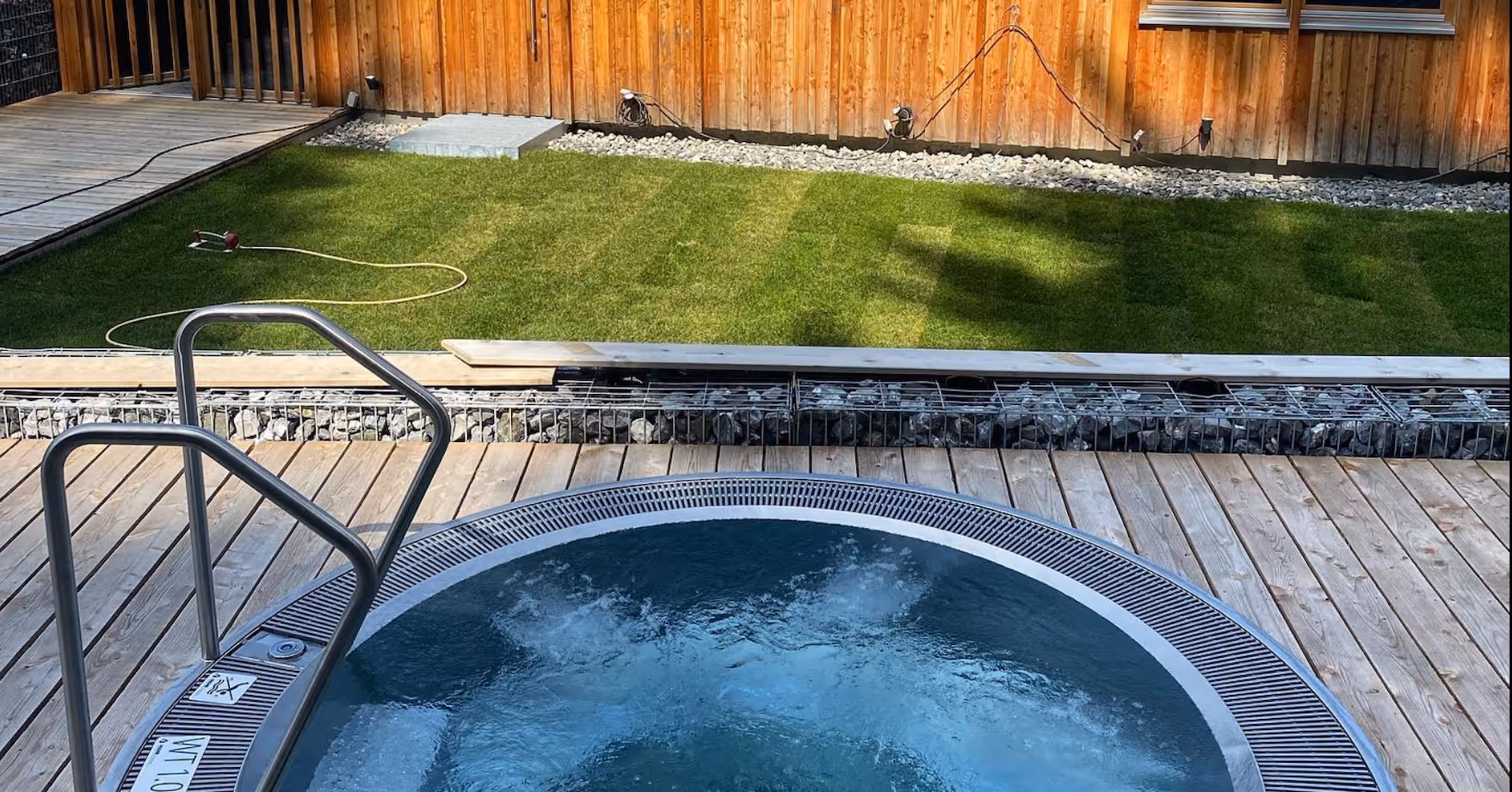 Blauer Whirlpool mit Wassersprudeln auf Holzdeck neben gepflegtem Garten mit grünem Rasen und Holzzaun.