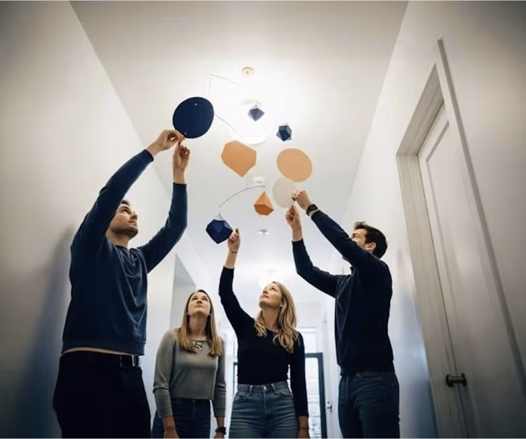 Four people, two men and two women, looking up and adjusting a mobile with various geometric shapes hanging from the ceiling.