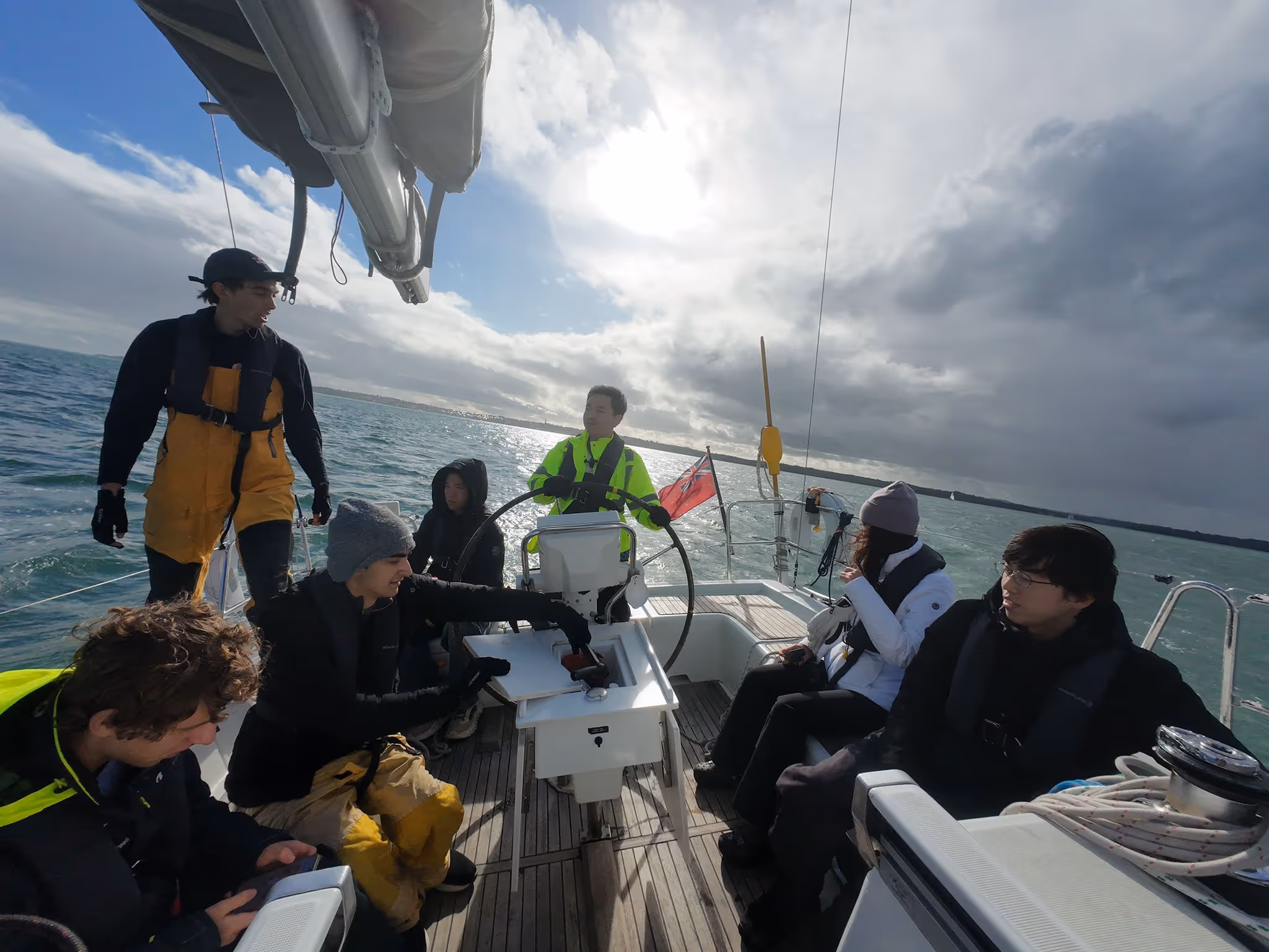 Group of seven people wearing life jackets sailing on a boat under a cloudy sky with bright sun reflecting on the water.