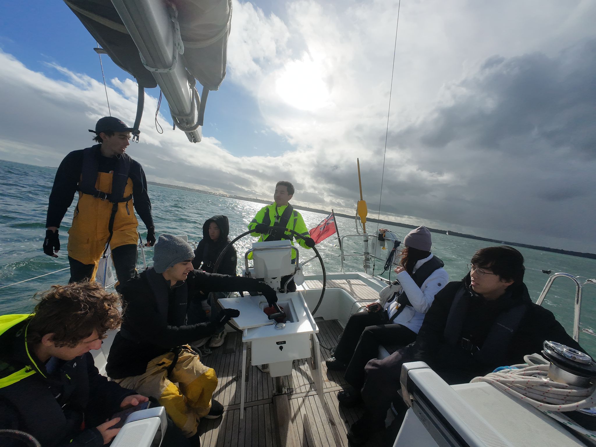 Group of seven people wearing life jackets sailing on a boat under a cloudy sky with bright sun reflecting on the water.
